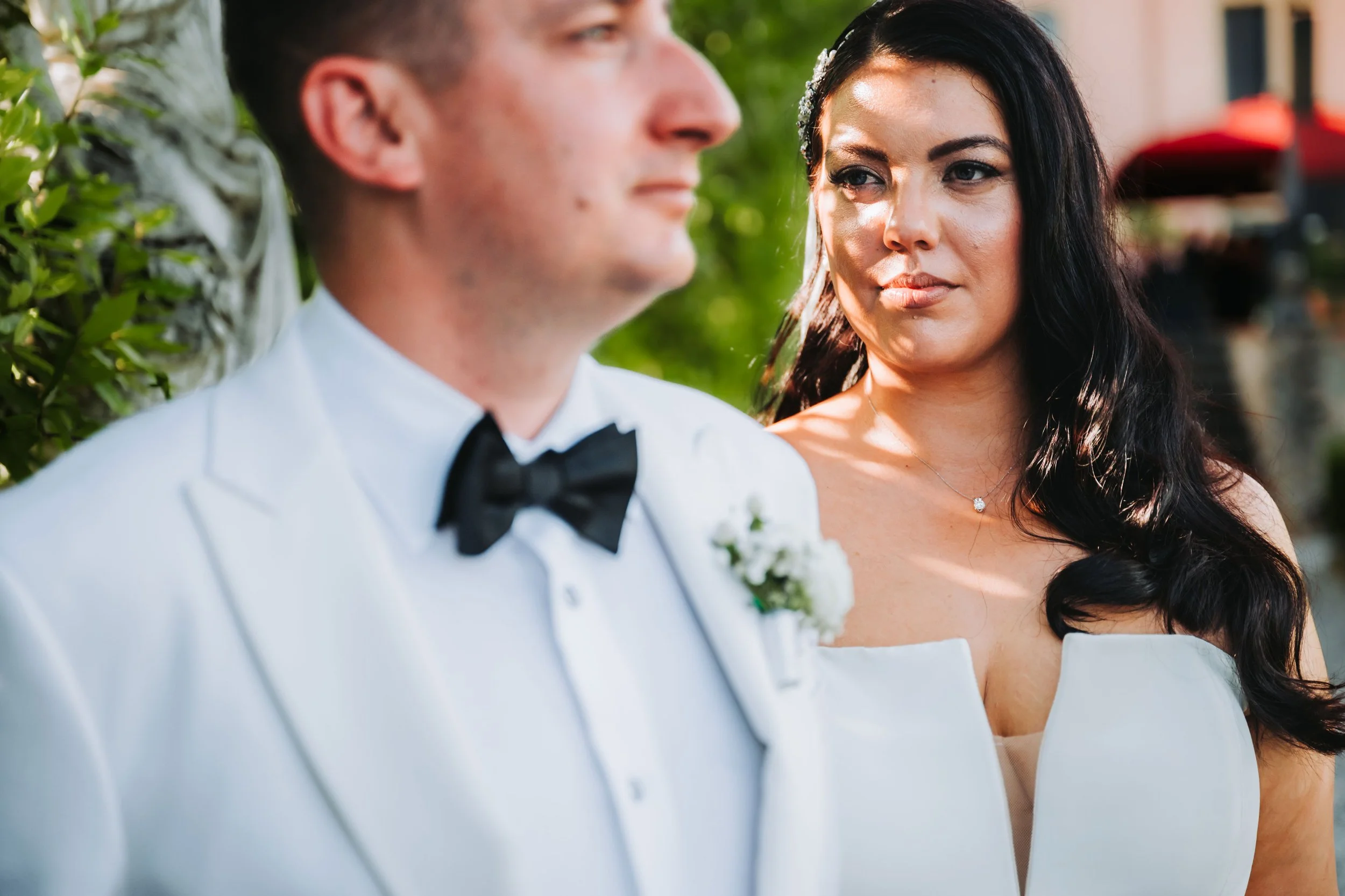 Una donna con capelli scuri e un vestito bianco osserva un uomo in abito da sposo con giacca bianca e papillon nero, ambientato all'aperto con luci soffuse e verde sullo sfondo.