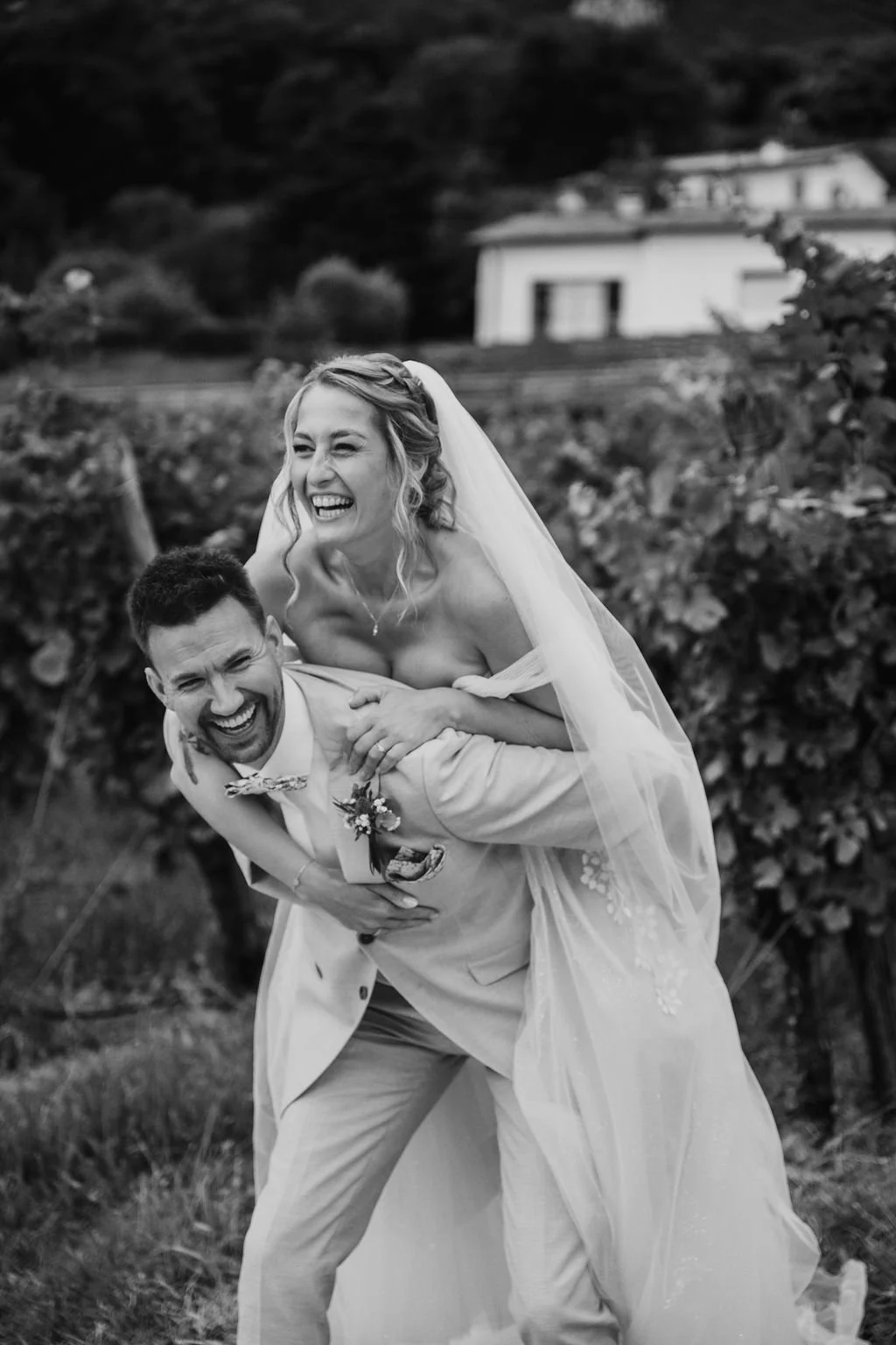 Una coppia di neo sposi sorridenti e felici, con la sposa sopra le spalle dello sposo, in un'ambientazione all'aperto, in bianco e nero.