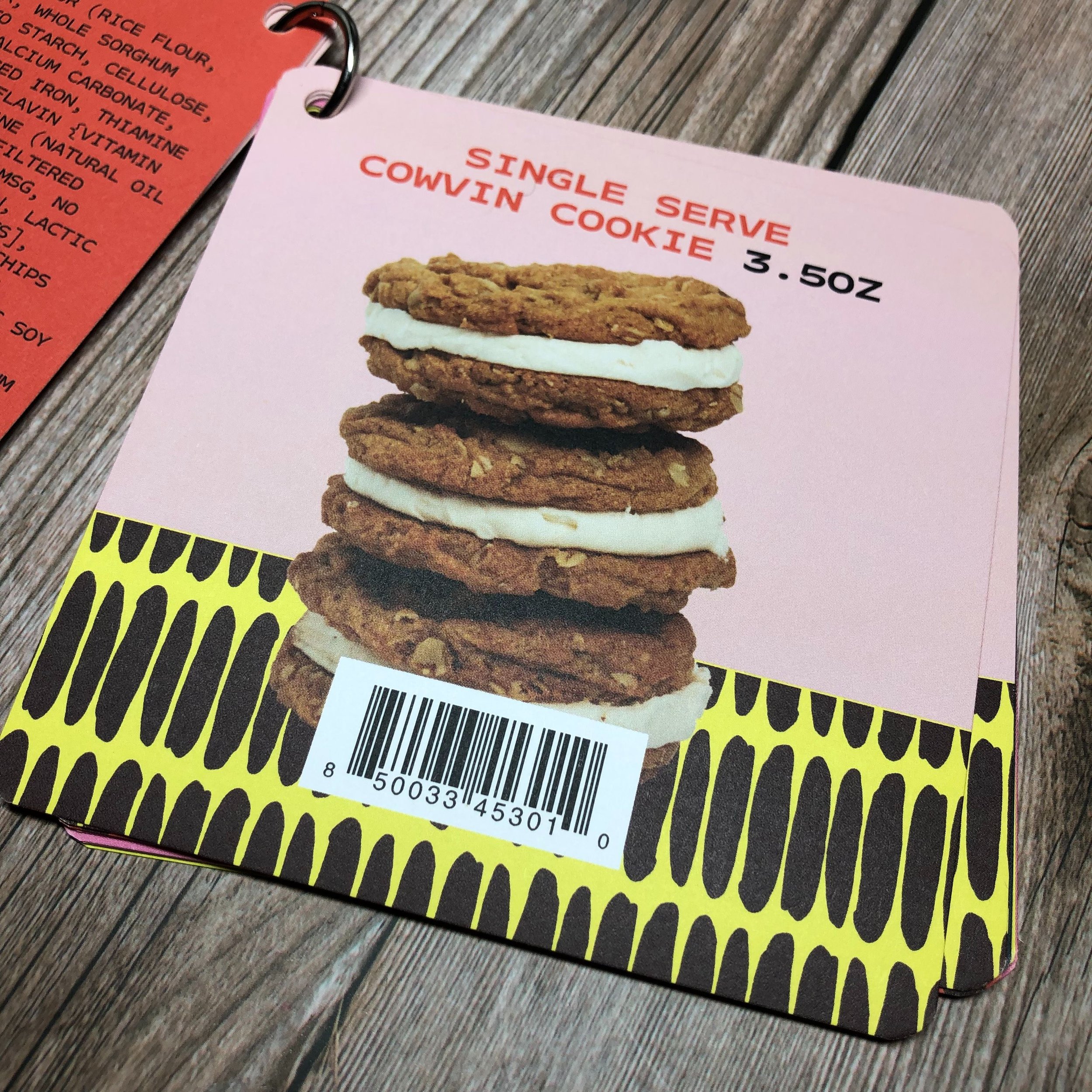 Pink menu page displaying a stack of four cookies with cream filling, labeled as 'Single Serve Cowin Cookie 3.5oz', with a barcode at the bottom and a yellow and black patterned border.