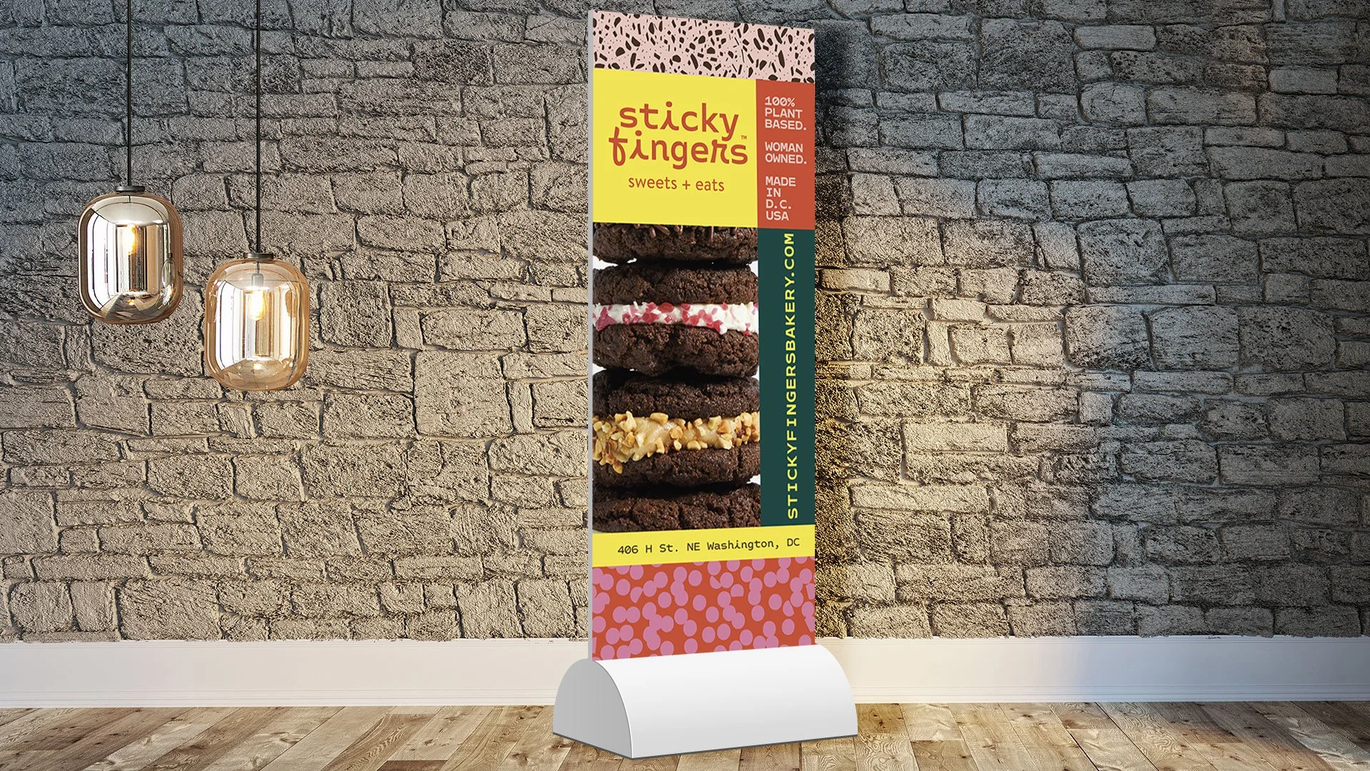 Vertical banner advertising Sticky Fingers sweets and eats, featuring images of layered cookies with various toppings, placed against a brick wall background with wooden floor, illuminated by two hanging pendant lights.