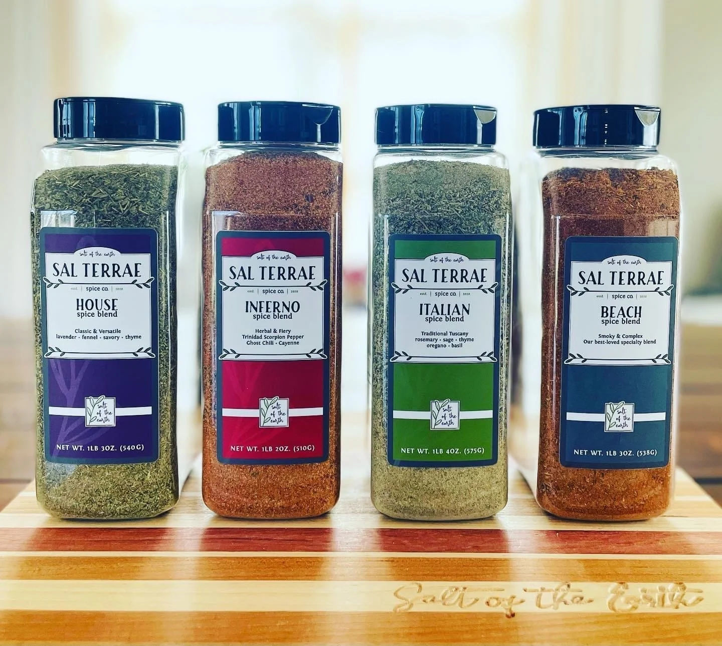Four spice jars labeled 'Sal Terrae' with different spice blends: House, Inferno, Italian, and Beach, placed on a wooden surface.