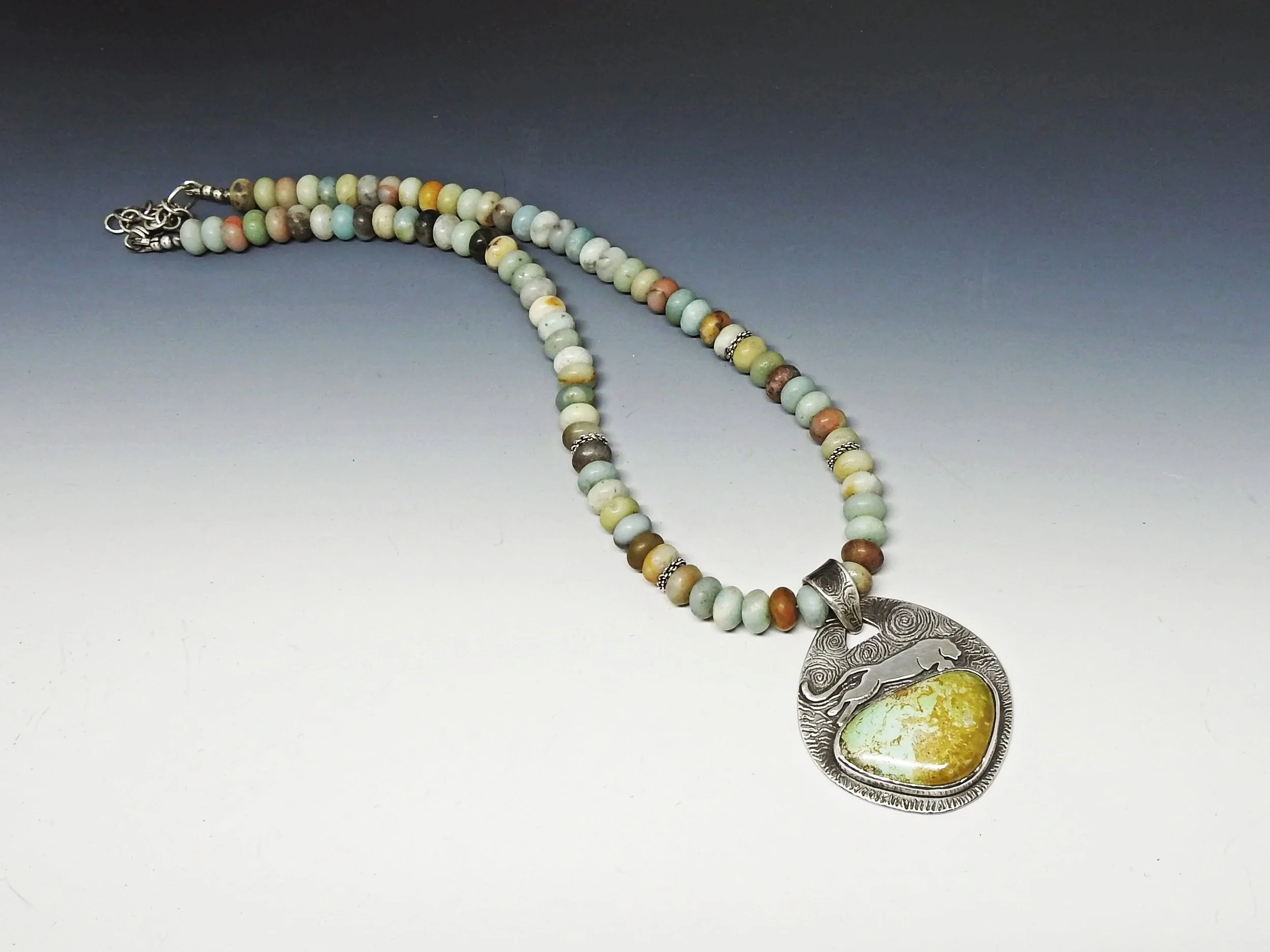 Beaded necklace with a large silver pendant featuring a green and yellow stone and a wolf design on a gray gradient background.