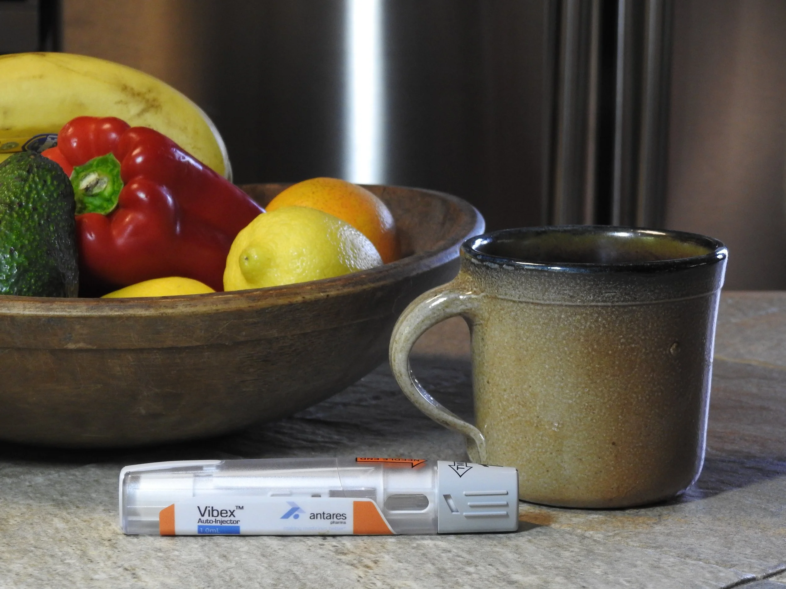 A ceramic mug, a vial labeled 'Vibex' vaccine, and a bowl of assorted fruits including bananas, red bell pepper, lemons, an orange, and an avocado on a textured surface.