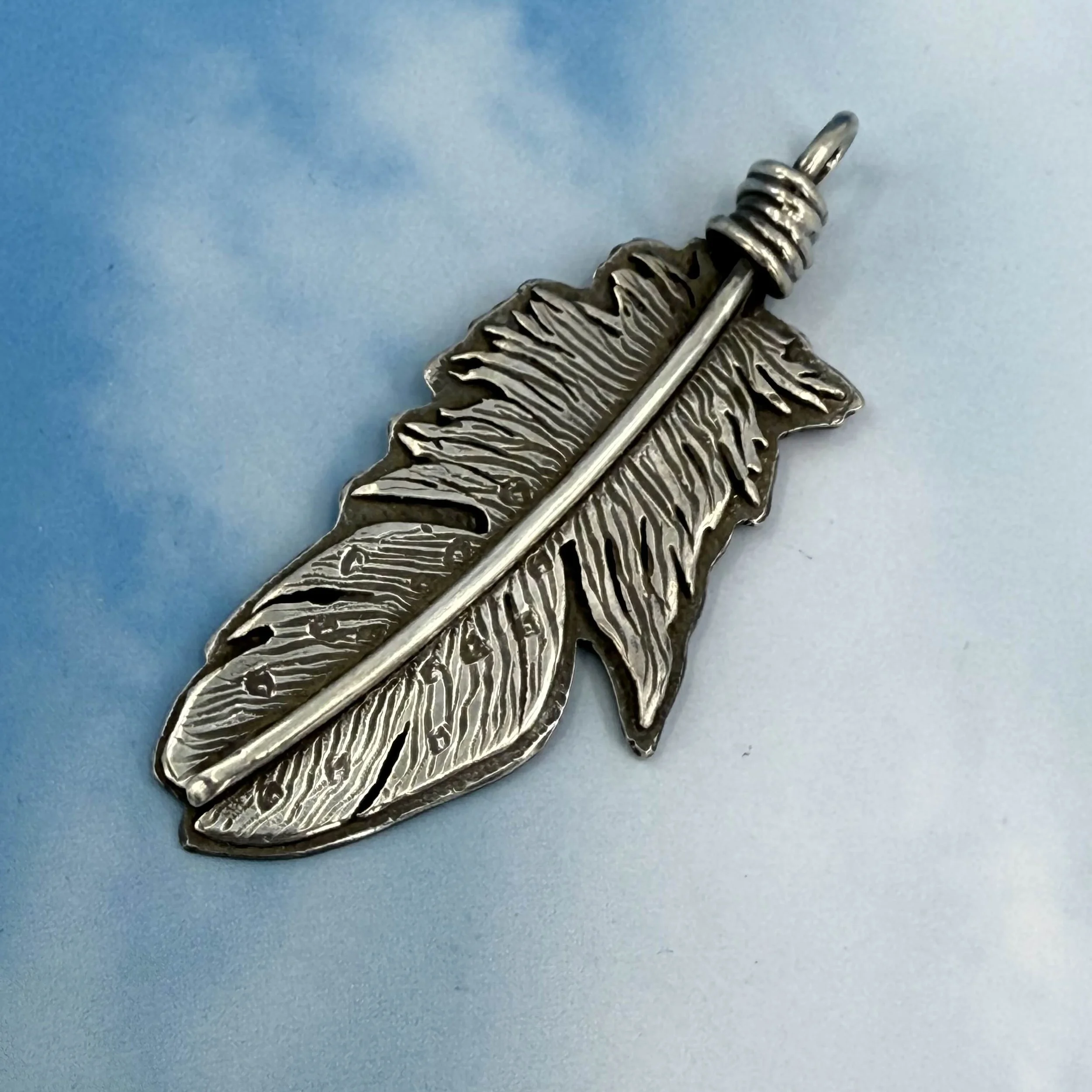 Silver feather-shaped pendant with a pin for jewelry, on a light blue background.