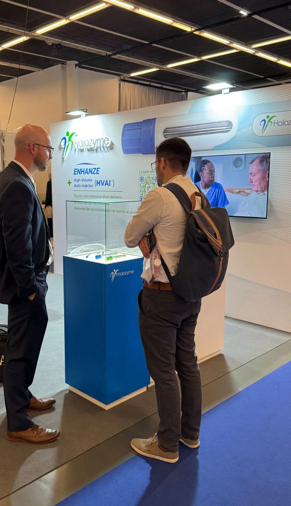 Two men are standing at a trade show booth with a large banner displaying the logo and product information for Halozyme, a pharmaceutical company. One man is gesturing toward a glass display case with medical devices, while the other man listens. In 
