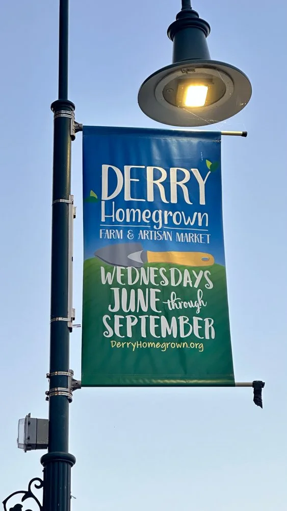 A street banner advertising Derry Homegrown Farm & Artisan Market, open Wednesdays from June through September, with a website link derryhomegrown.org, attached to a street lamp with an overhead light.