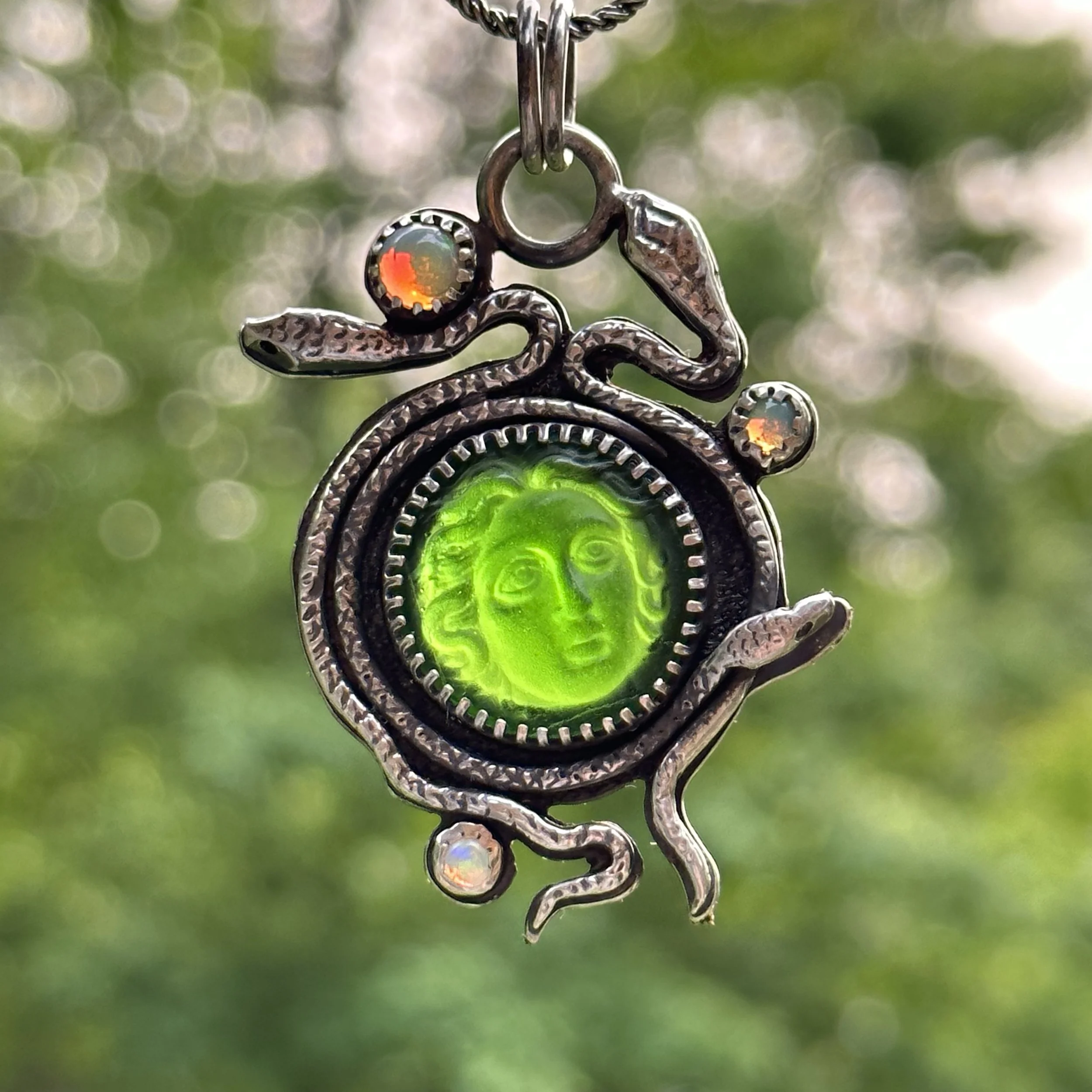 A silver pendant with a green glass centerpiece depicting a face, wrapped by a snake design with small gemstones, hanging against a green blurred background.
