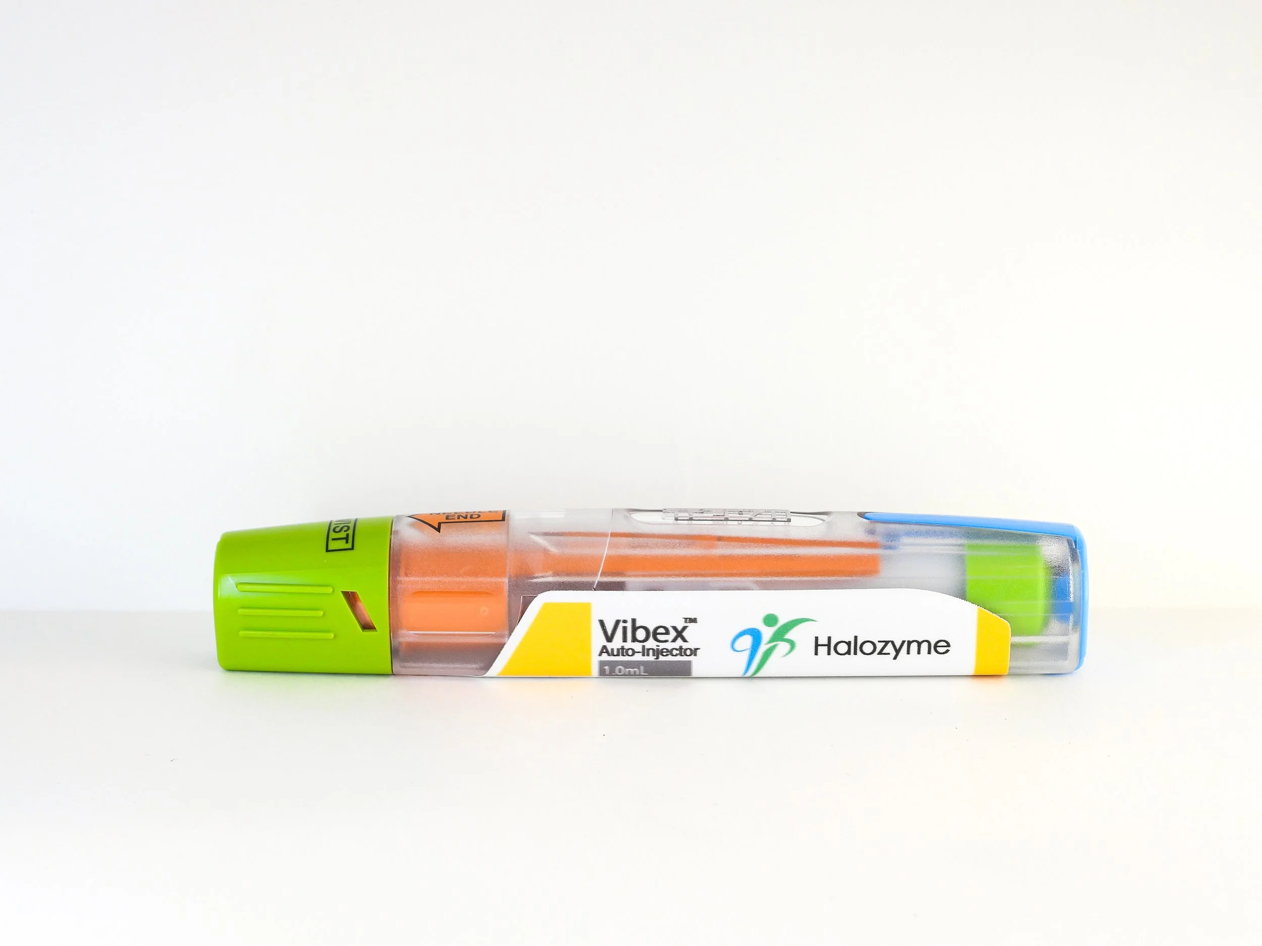 A medical auto-injector pen labeled 'Vibex™ Auto-Injector' containing Halozyme, with a green cap and orange needle cap, lying on a white surface against a plain white background.