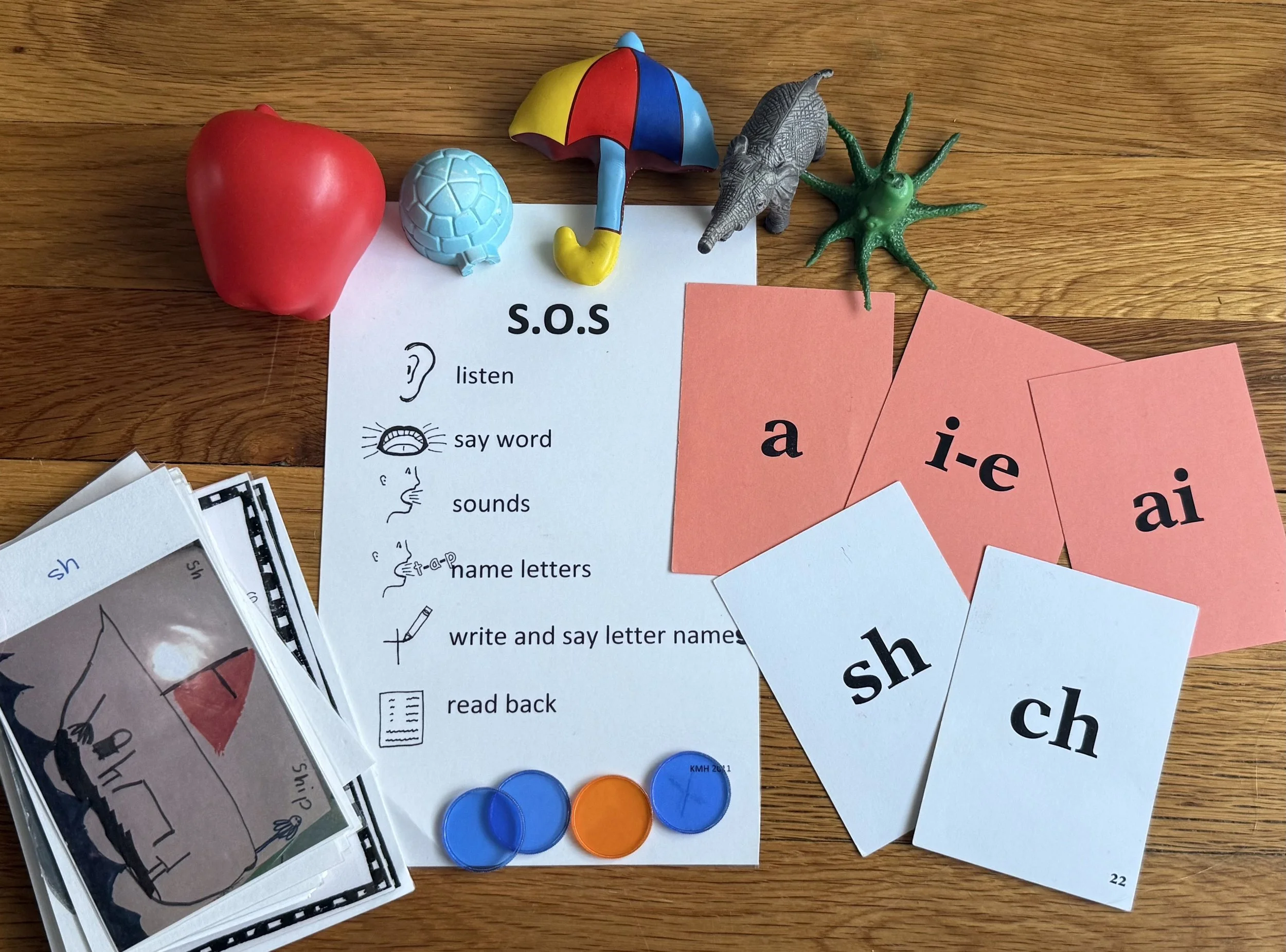 Educational materials for teaching spelling and phonics, including flashcards with 'sh', 'ch', 'a', 'i-e', 'ai', a printed chart with SOS instructions, small toy objects like an apple, turtle, umbrella, whale, and octopus, and a stack of cards in the corner.
