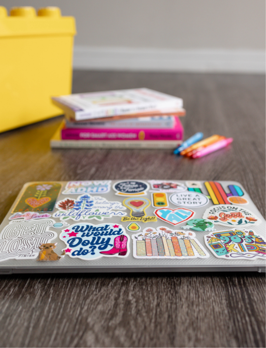 A closed laptop decorated with colorful stickers on a wooden surface, with a stack of notebooks or planners and three colored pens nearby, and a yellow storage bin in the background.