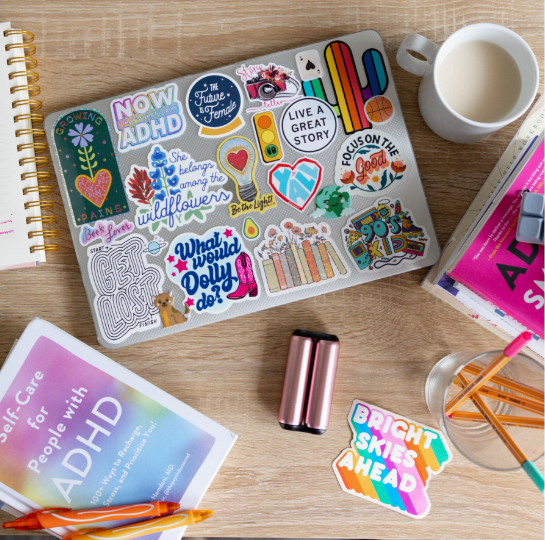 Laptop decorated with colorful stickers in a workspace with notebooks, a cup of milk, highlighters, and a self-care book.