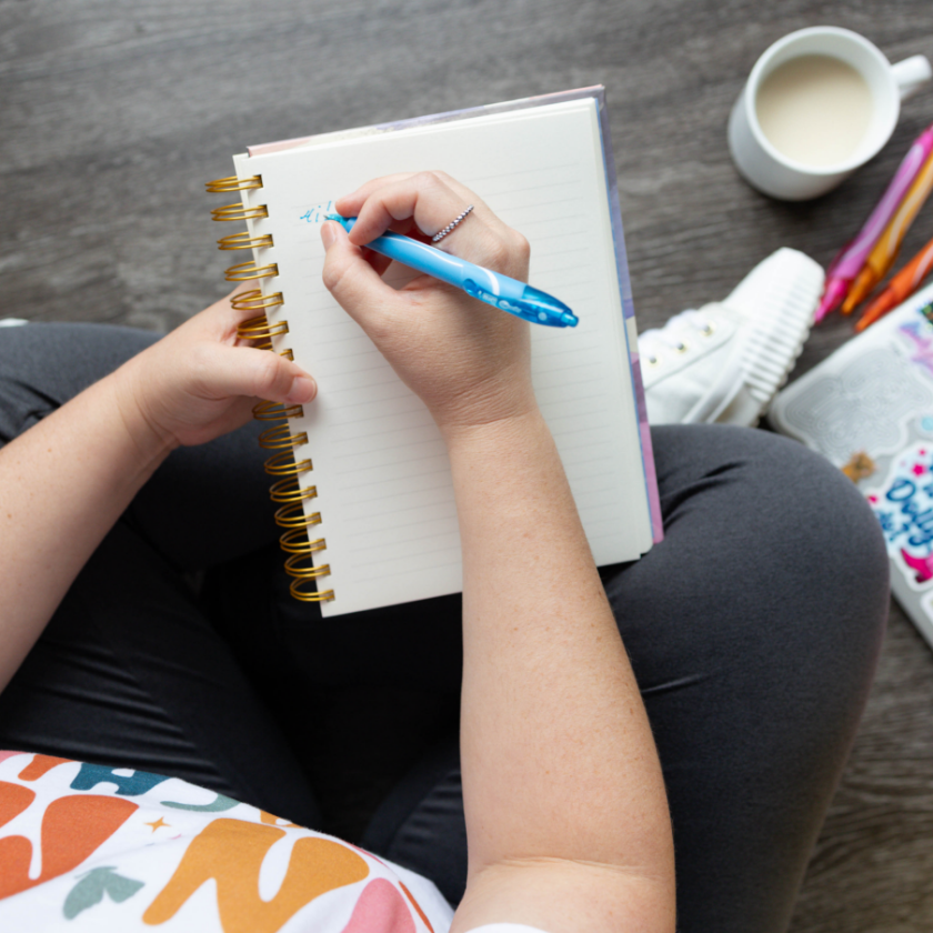 An overhead view of a person writing in a spiral notebook with a blue pen, sitting on their lap. There is a mug of milk or cream, sneakers, and other stationery nearby on a wooden floor.