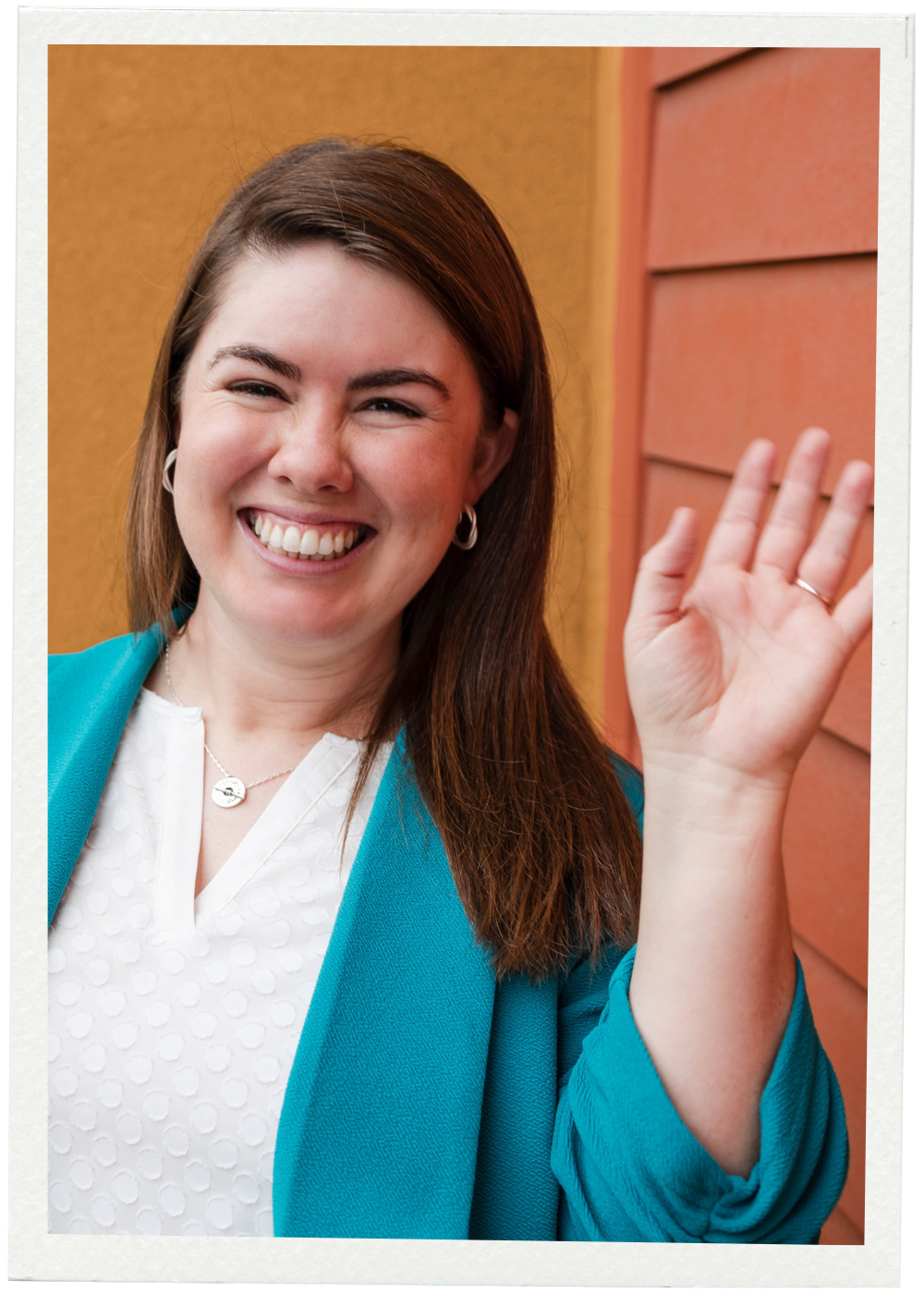 A smiling woman with brown hair wearing a white top and turquoise blazer, waving against a background with a brown-orange wall.