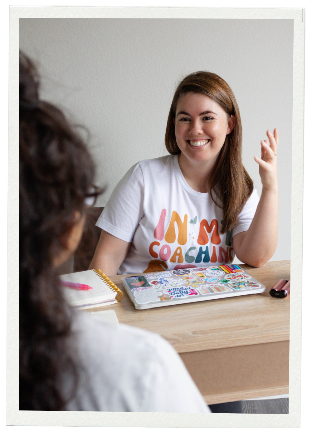 A woman with long brown hair smiling and gesturing with her hand while talking to another person during an interview or discussion, seated at a desk with a laptop covered in stickers, a notebook, and a pink pen.