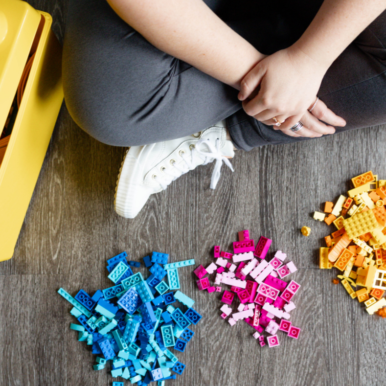 Person sitting on the floor with legs crossed, wearing white sneakers and gray pants, playing with colorful LEGO bricks on a wooden floor.