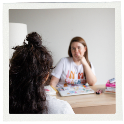 A woman with curly dark hair sitting across from a woman with straight light brown hair at a table, having a conversation in a room with white walls.