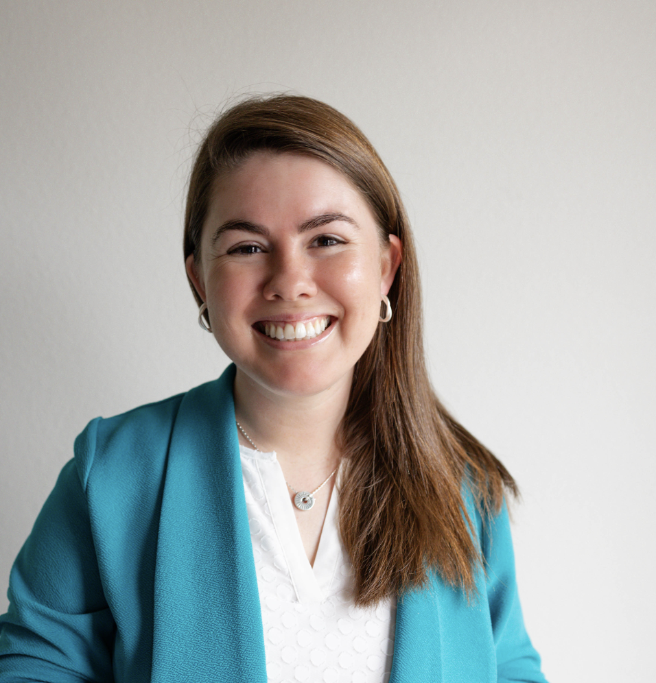 A woman with shoulder-length brown hair, smiling, wearing a teal blazer and white top, against a plain neutral background.