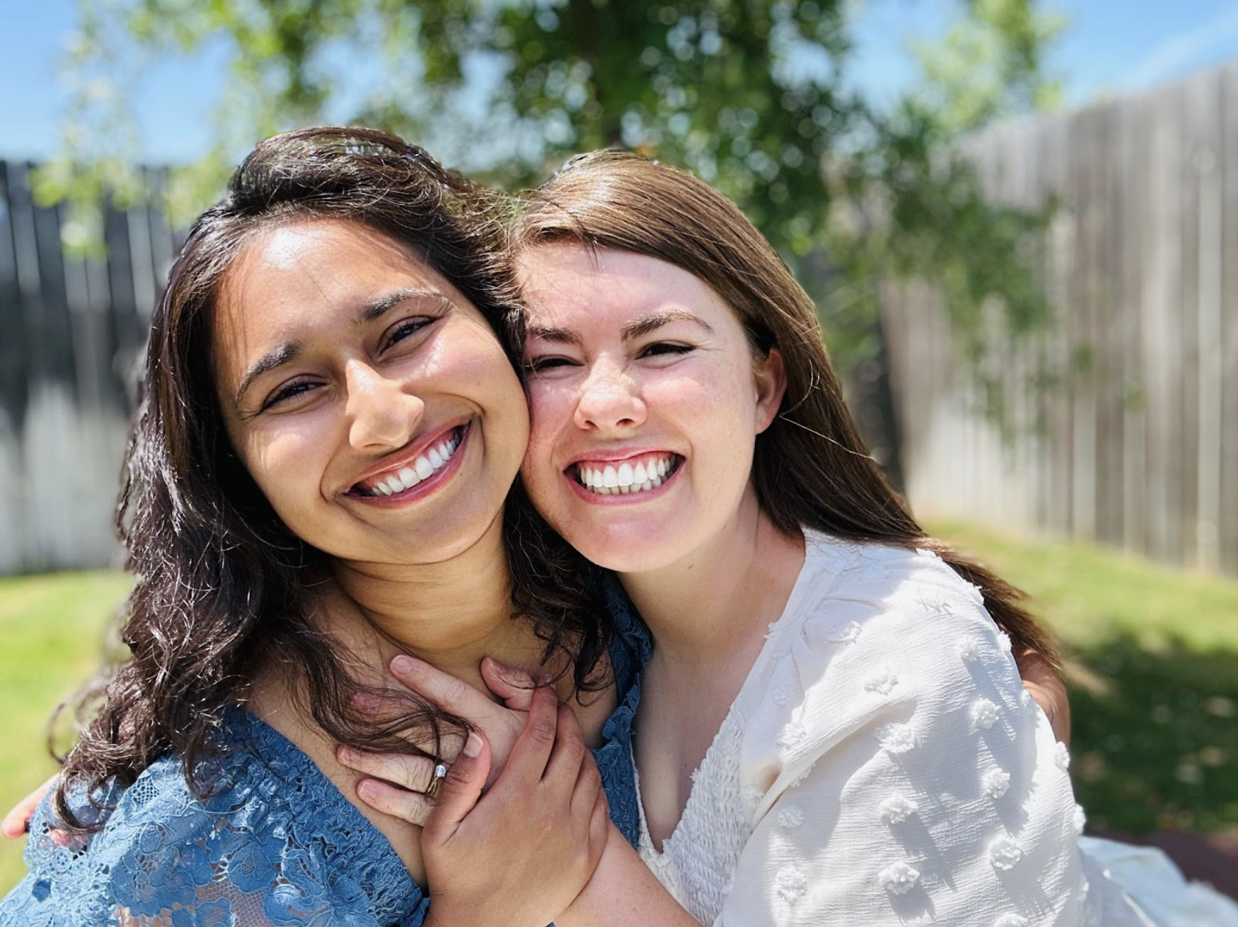 Two women smiling and hugging outdoors in a sunny backyard with trees and a wooden fence.