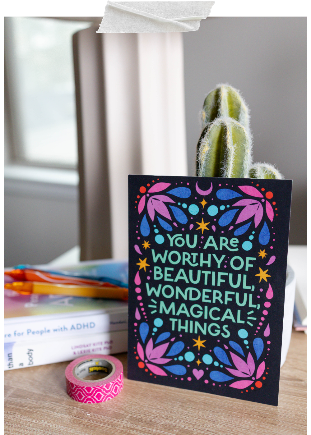 A colorful motivational card with purple, blue, pink, yellow, and green designs and the words "You are worthy of beautiful, wonderful, magical things" stands upright on a wooden surface. Behind it is a cactus plant with fuzzy white spines, and to the left are books, pens, and pink decorative tape near a window with blinds.