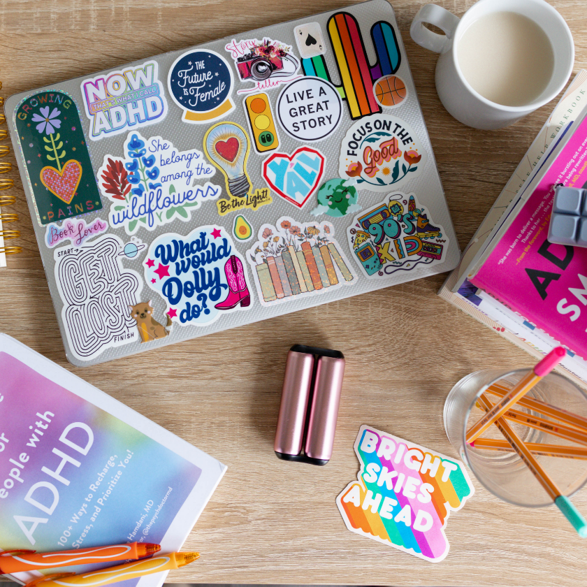 A desk with a laptop covered in colorful stickers, a white mug filled with milk or coffee, a transparent glass with pencils, a book, some papers, a pink battery, and a pink marker.