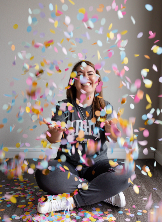 A young woman sitting on the floor, smiling with eyes closed as colorful confetti falls around her.
