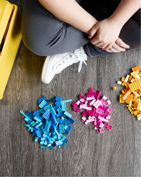 Person sitting on the floor with legs crossed, wearing white sneakers and gray pants, surrounded by colorful toy building blocks arranged in separate piles of blue, pink, and yellow.