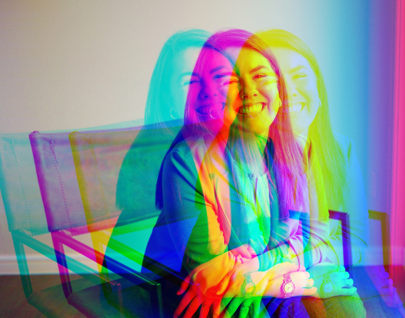 A woman sitting on a chair smiling, with vibrant color overlays creating a layered, rainbow effect.