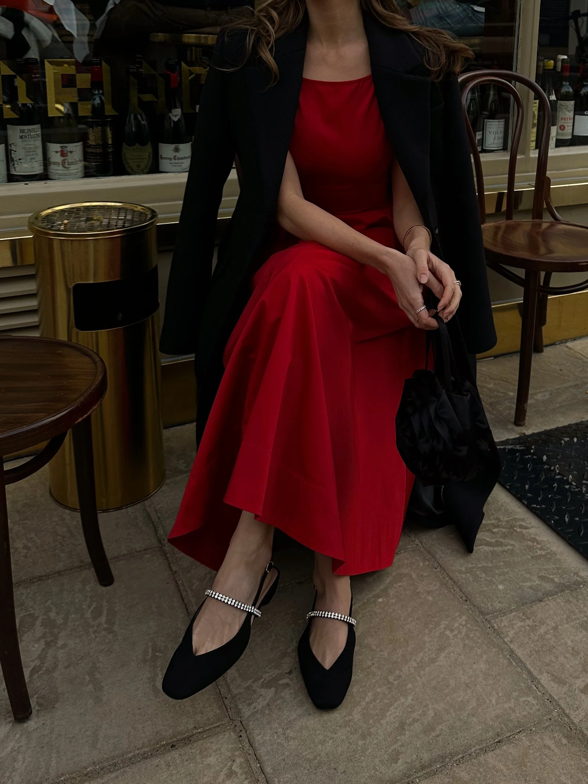 Woman sitting outside wearing a red dress, black coat, and black shoes with rhinestone straps. Holding a black purse.