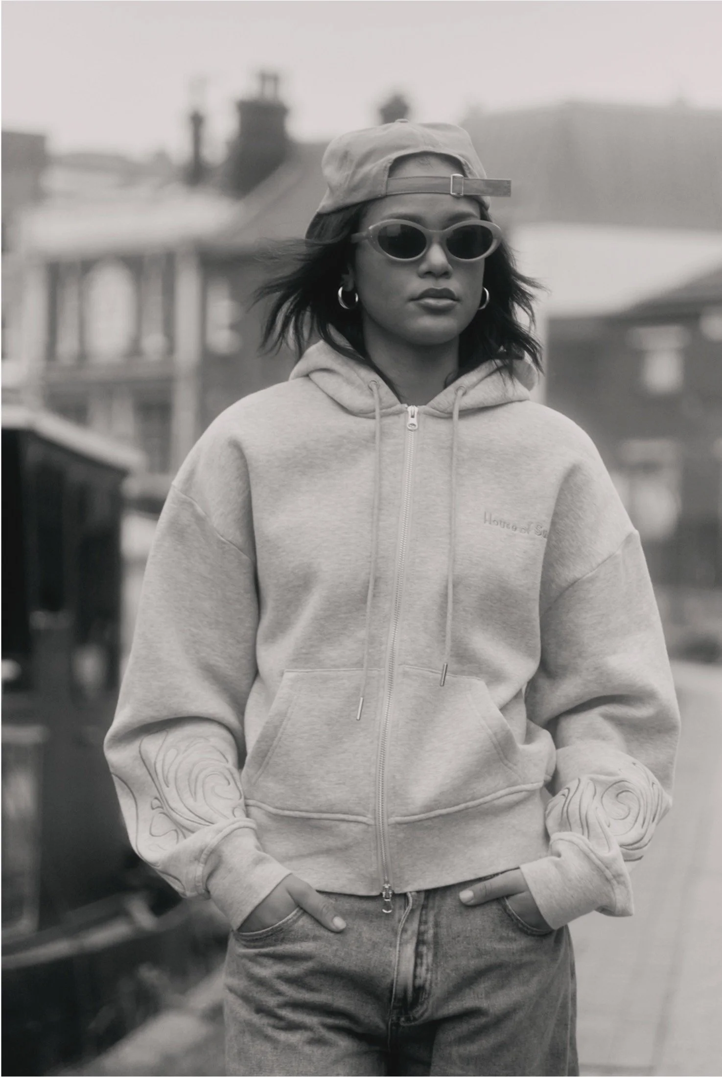 Person wearing a hoodie, sunglasses, and a backward cap in an urban setting.