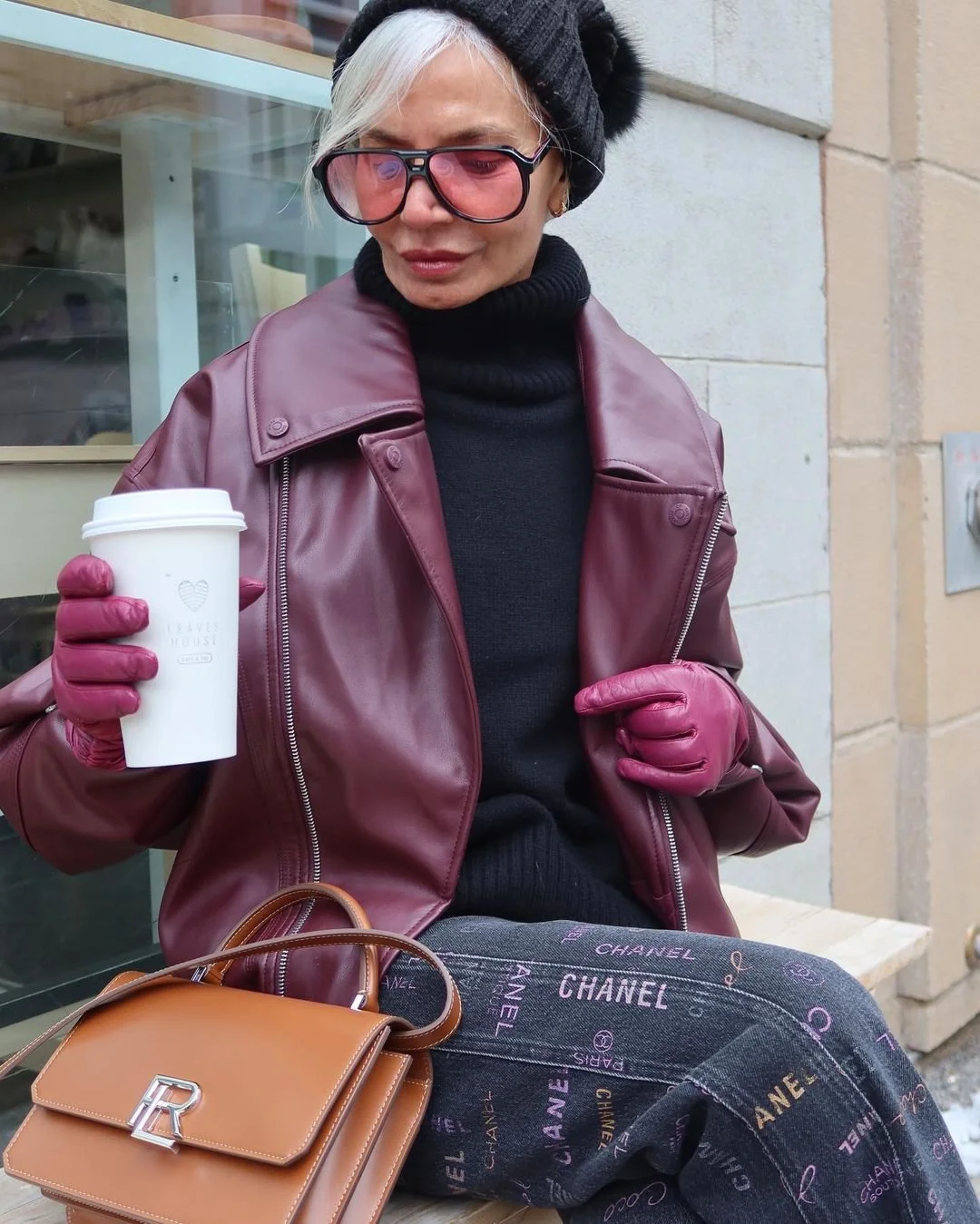 Stylish person wearing maroon leather jacket, pink gloves, black turtleneck, grey patterned pants, holding a coffee cup, and carrying a brown handbag with initials "R."