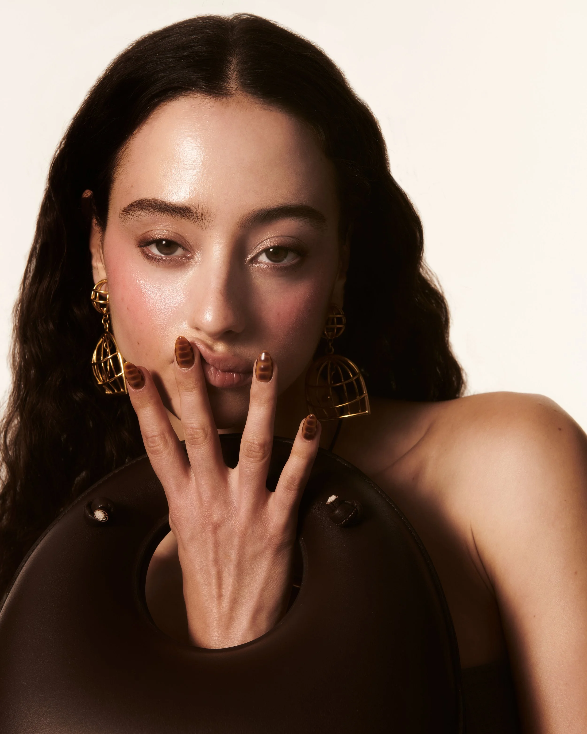 Woman with long dark hair wearing gold earrings, holding a brown circular object, with fingers touching her lips, and displaying brown nail art.