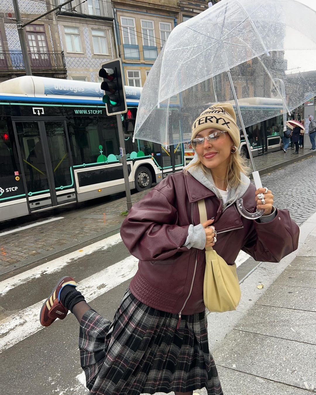A person standing on a street crossing holding a clear umbrella, wearing a brown jacket, plaid skirt, and sneakers, with a beanie. City bus and buildings in the background.