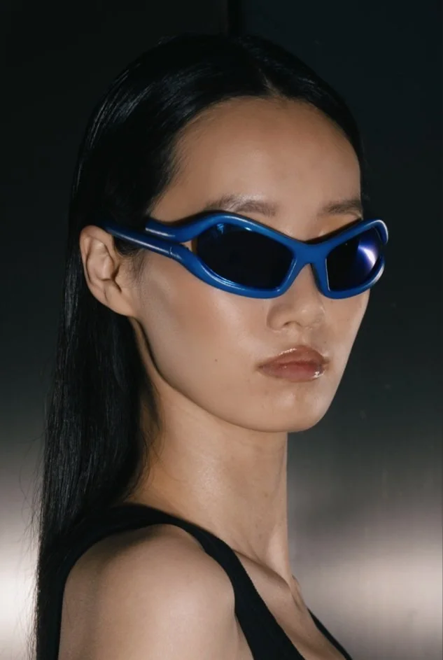 Person wearing blue sunglasses, black hair, with a neutral expression, against a dark background.