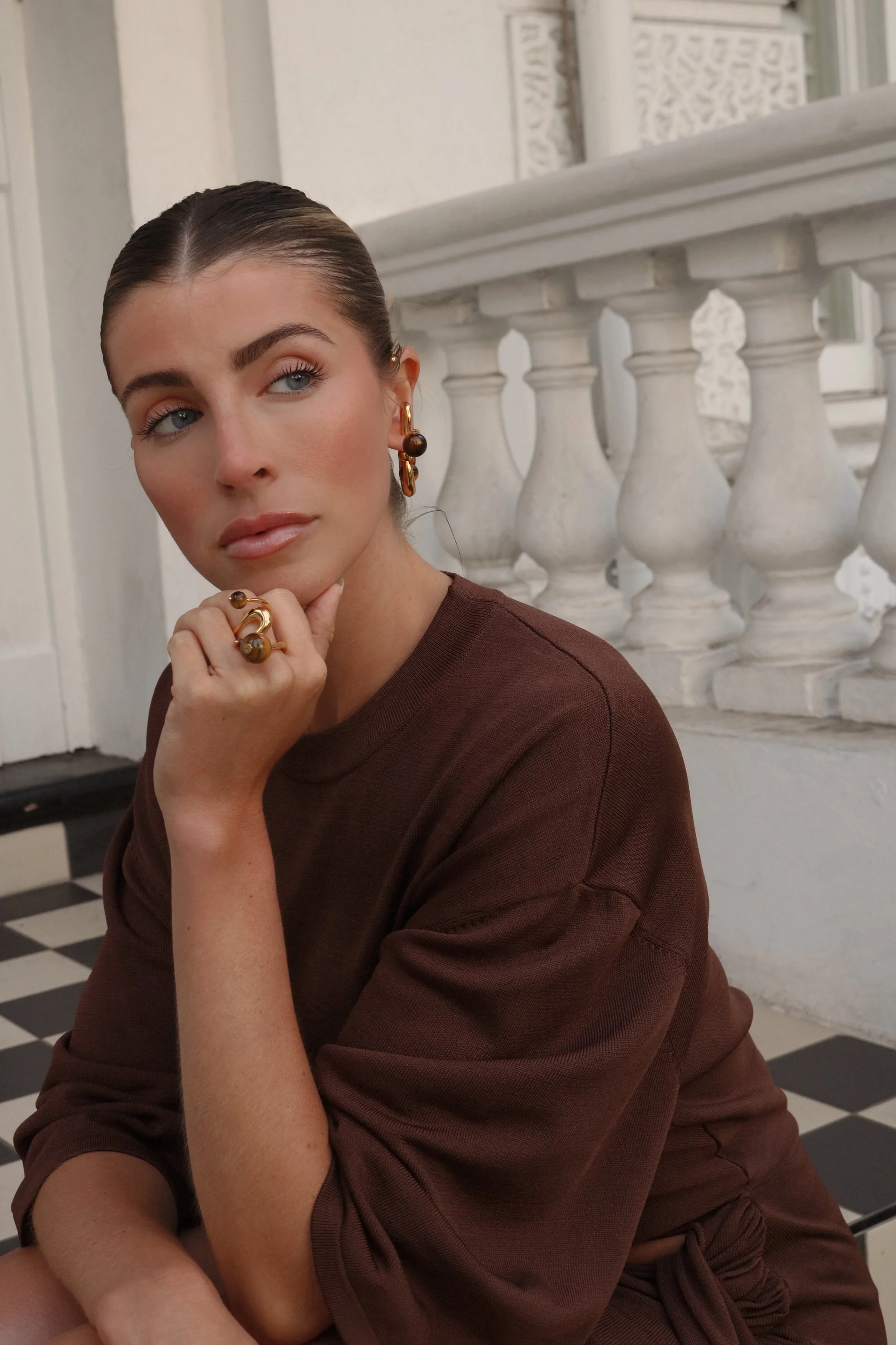 Person wearing brown outfit, gold jewelry, sitting on checkered floor near white balustrade.