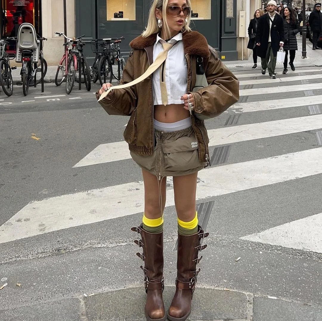 Fashionable person wearing a brown jacket, cropped white shirt, tie, short khaki skirt, tall brown boots, and yellow socks, standing on a city street with bicycles in the background.