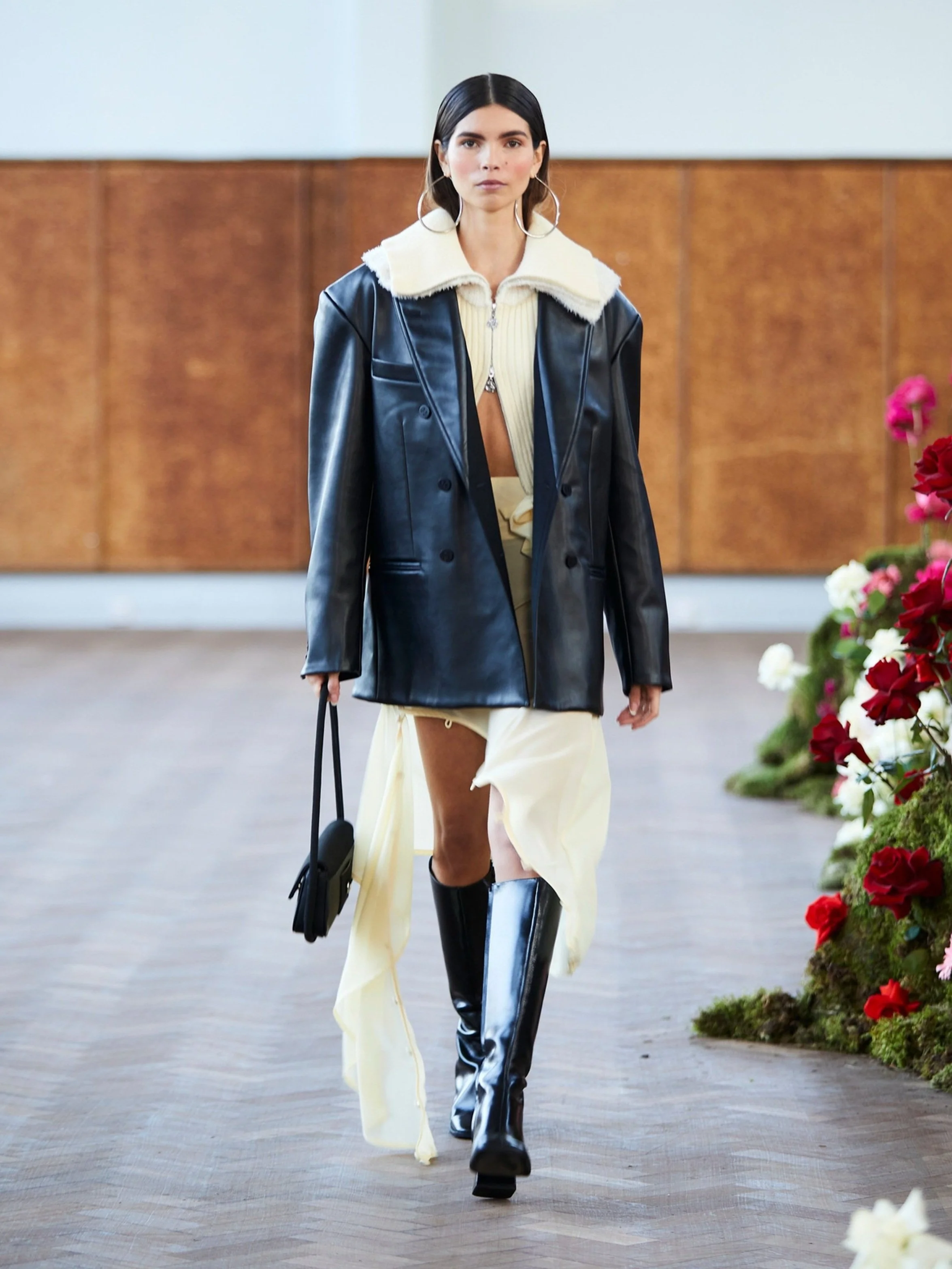 Fashion model wearing a black leather coat with a cream collar, cream dress, and knee-high black boots on a runway.