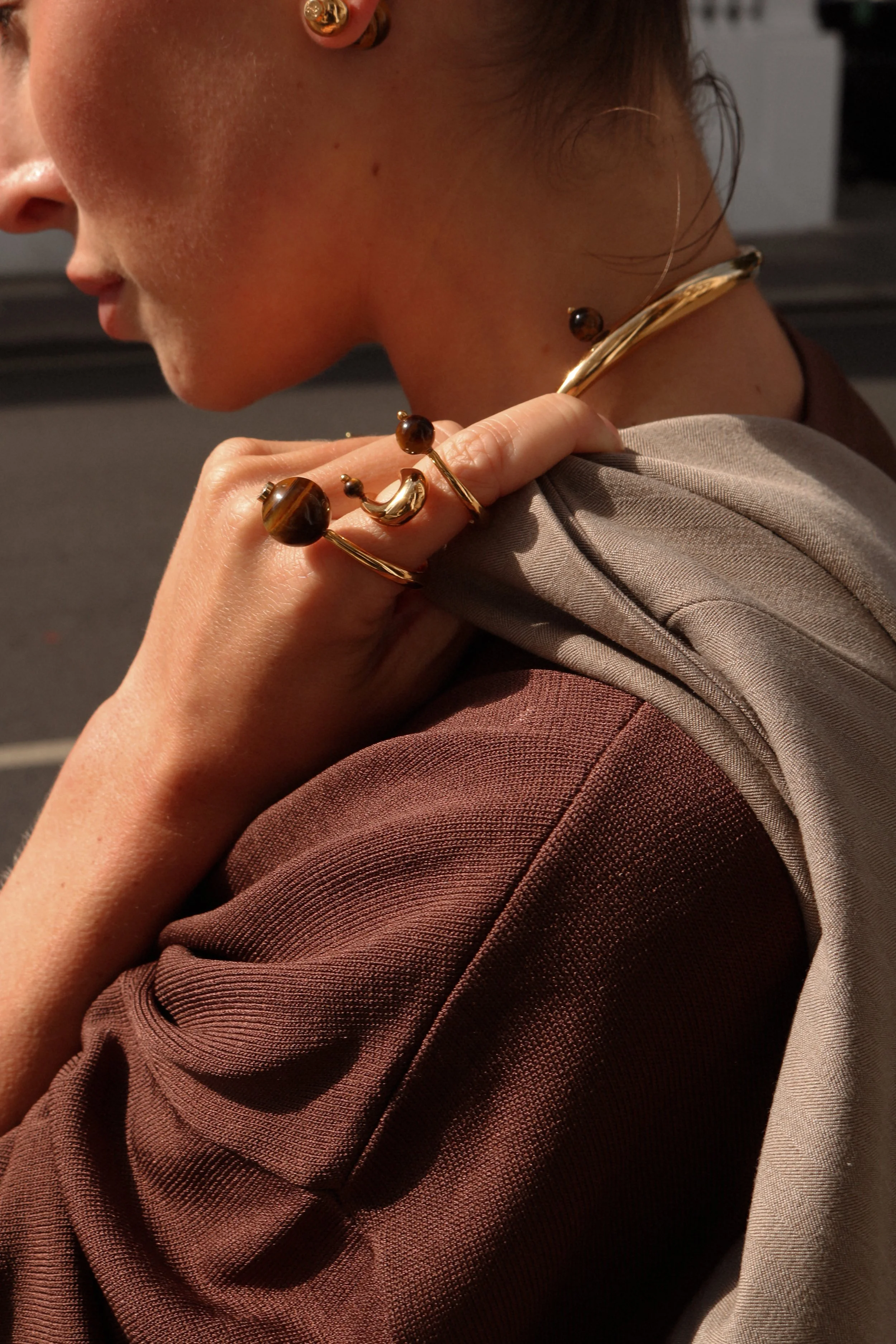 Person wearing gold jewelry with brown gemstones, including earrings, rings, and a bracelet, with a beige and brown outfit.