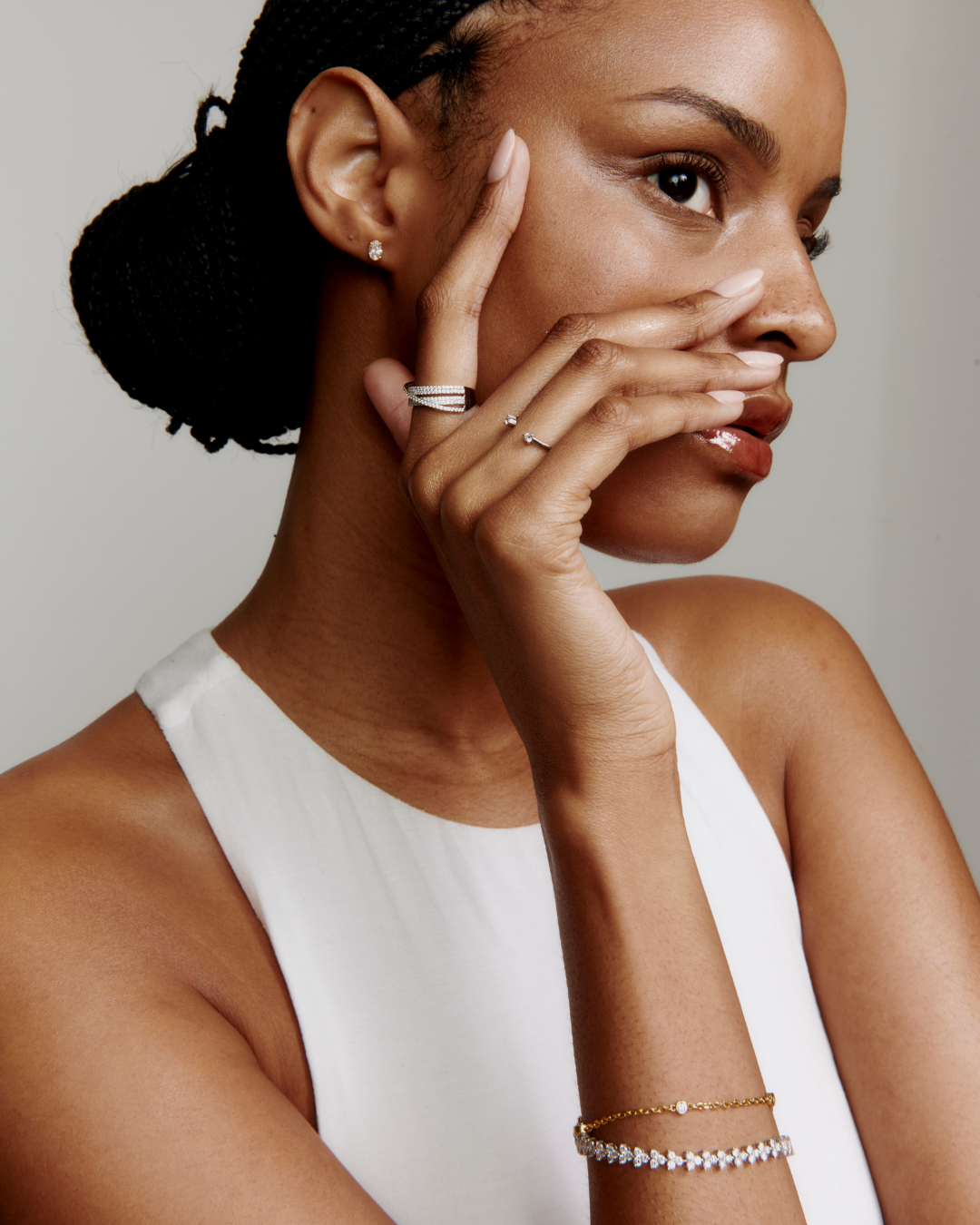 Woman wearing jewelry including earrings, rings, and bracelets, with braided hair, wearing a white sleeveless top against a neutral background.