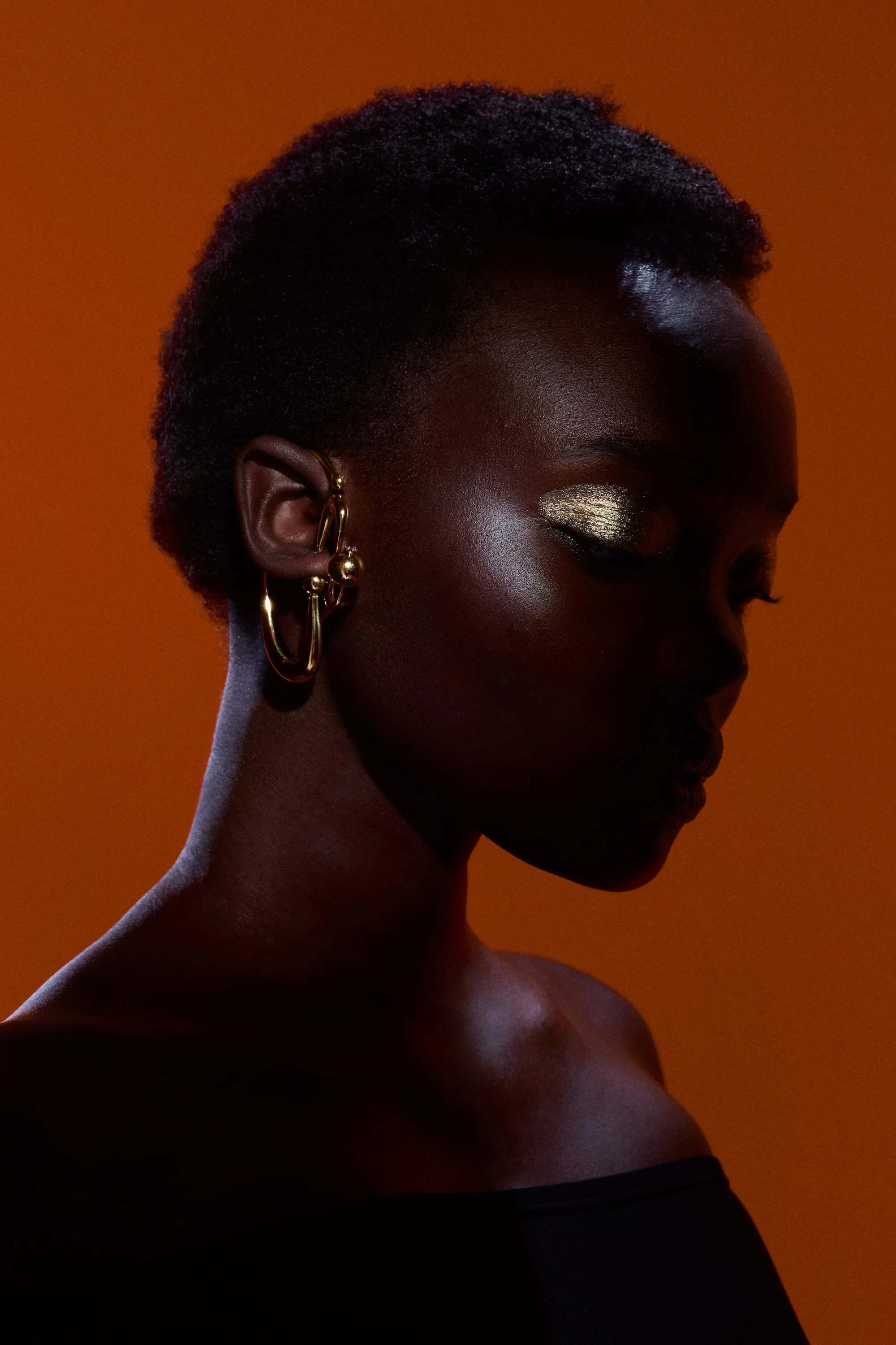 Silhouette of a person with short hair, wearing gold earrings, and gold eyeshadow, against an orange background.
