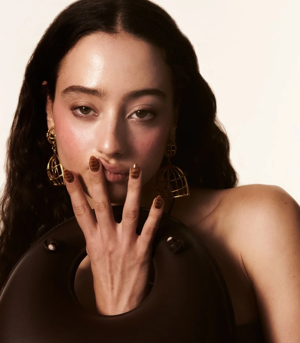 Woman with long hair, holding a brown circular handbag, wearing gold geometric earrings, brown patterned nails, and minimal makeup.