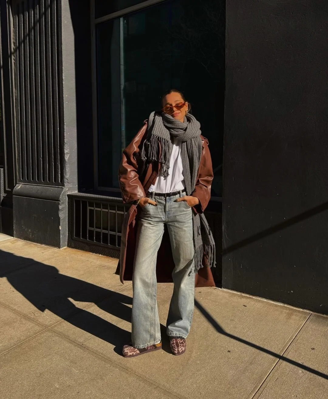 Person wearing a brown leather coat, gray scarf, and sunglasses, standing on a city sidewalk.
