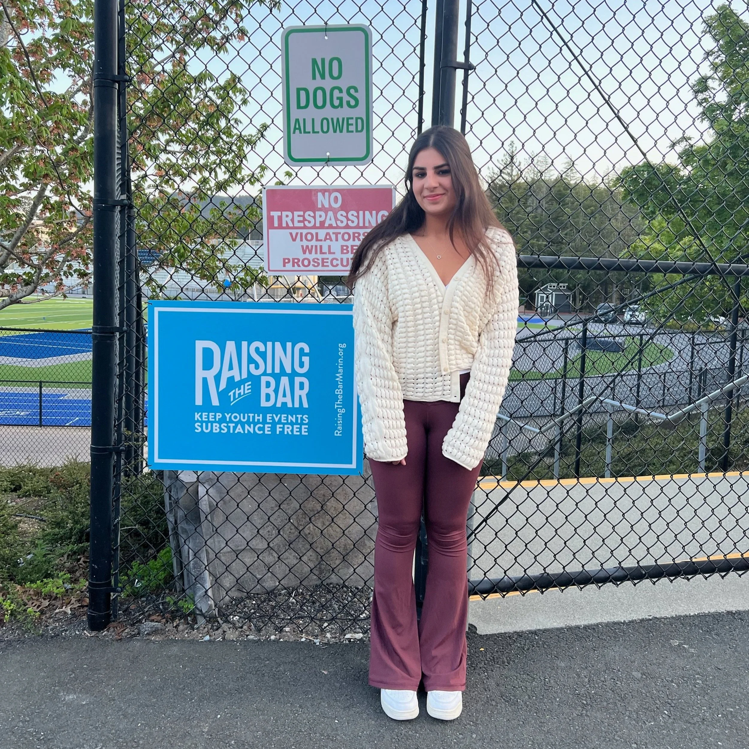 A young woman standing in front of a chain-link fence with signs that read "No Dogs Allowed", "No Trespassing Violators Will Be Prosecuted", and a blue sign for raising the bar and keeping youth events substance free. She is wearing a white knitted sweater, maroon bell-bottom pants, and white shoes, with a baseball field visible in the background.