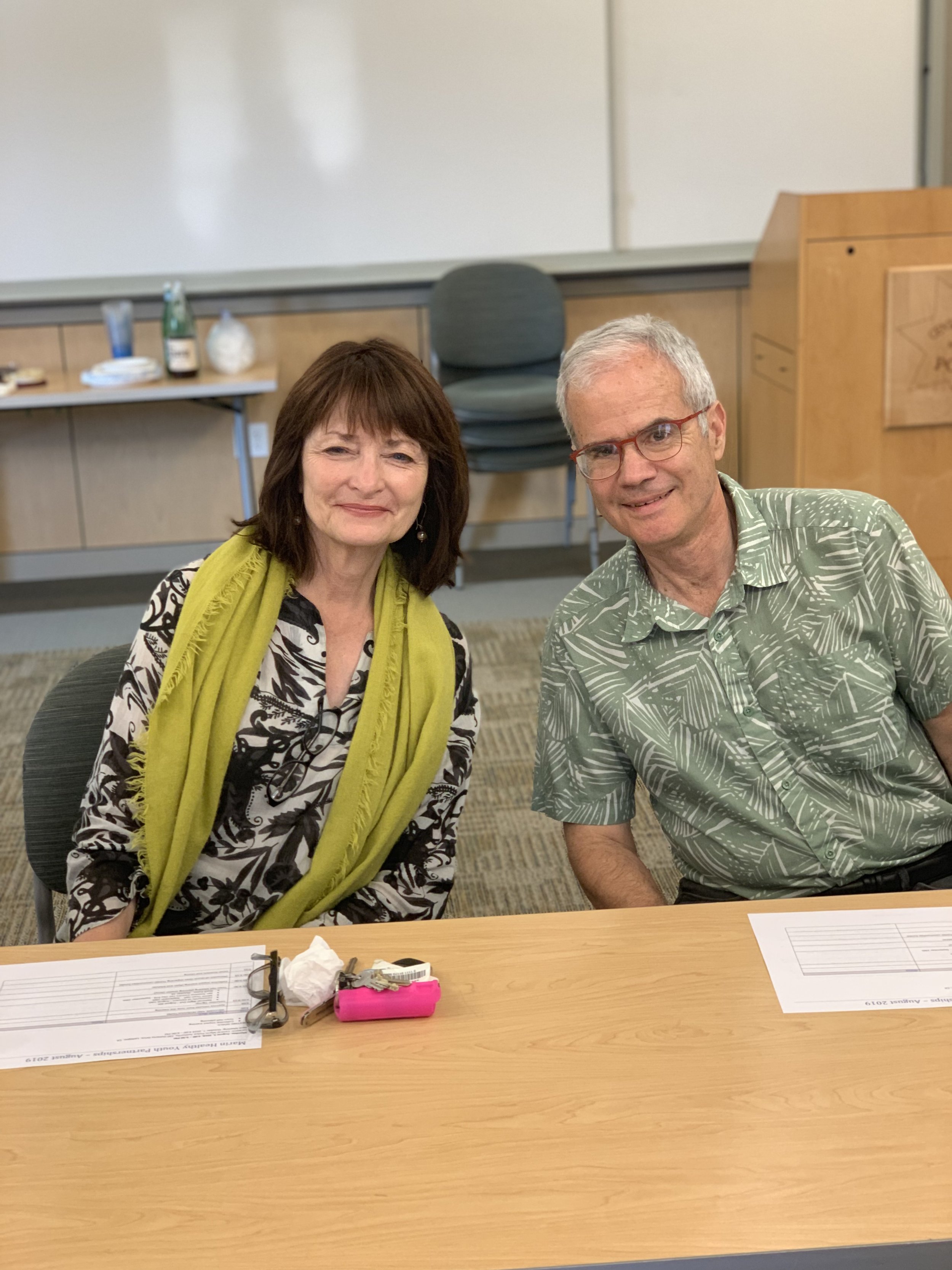A woman with shoulder-length brown hair, dressed in a black and white patterned blouse with a yellow scarf, seated next to a man with short white hair, glasses with red frames, wearing a green tropical-themed shirt, sitting at a table in a classroom or conference room.