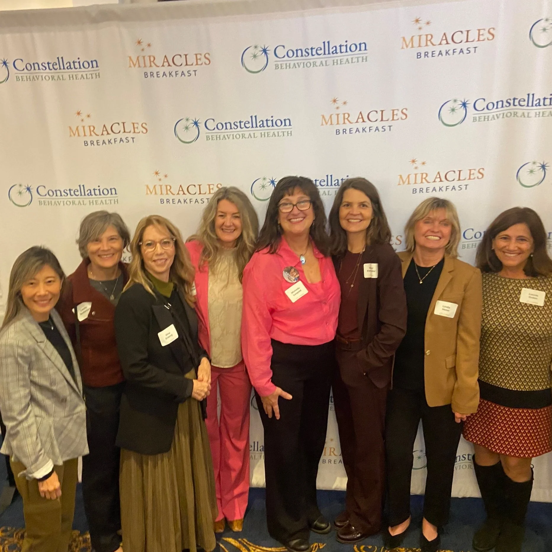 Group of women standing together at an event, posing in front of a backdrop with logos for Constellation Behavioral Health and Miracles Breakfast.