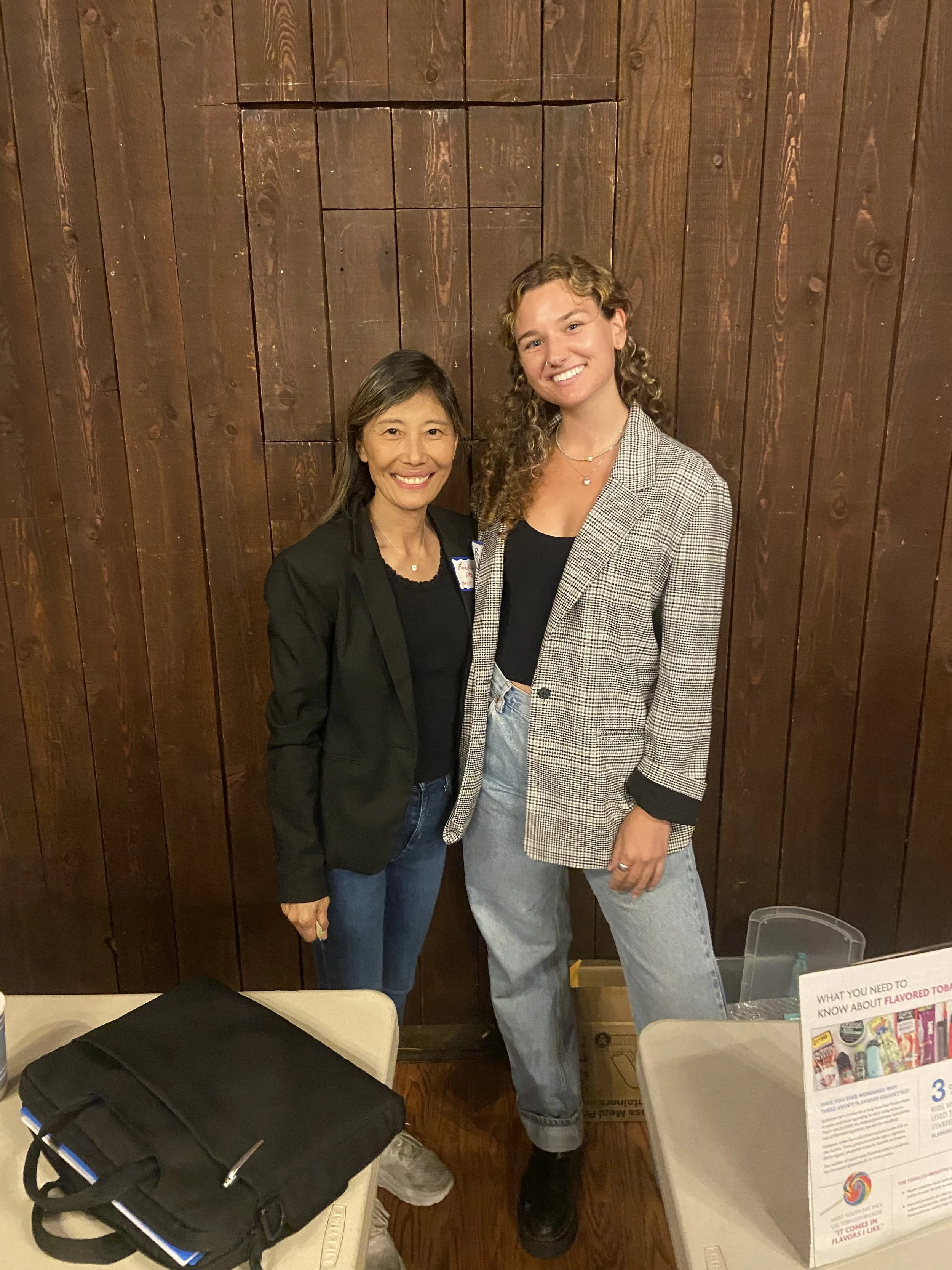 Two women standing close and smiling in front of wooden wall, one in a black blazer and the other in a plaid blazer and jeans.