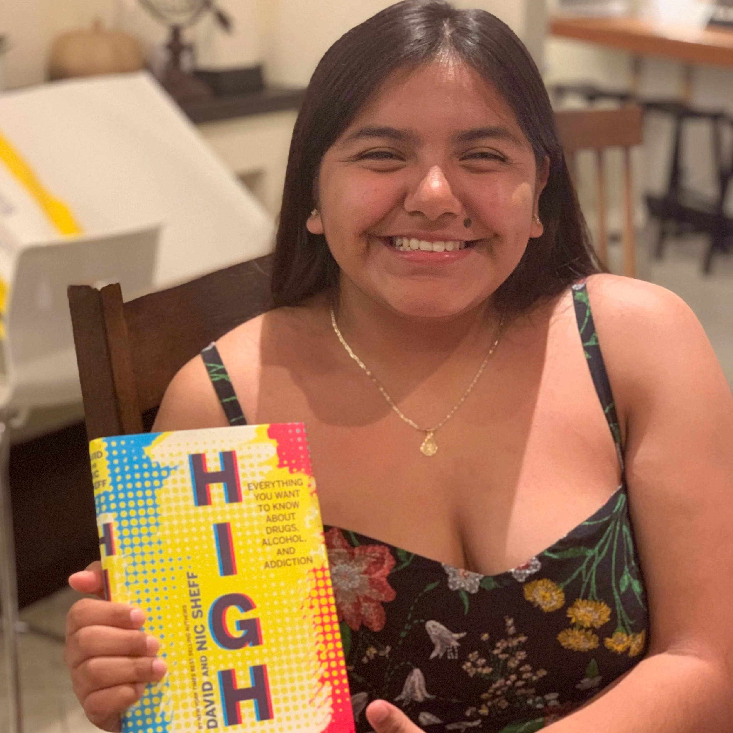 A young woman with dark hair pulled back, wearing a floral dress and a gold necklace, smiling and holding a colorful book titled "High" at a dining table in a home setting.