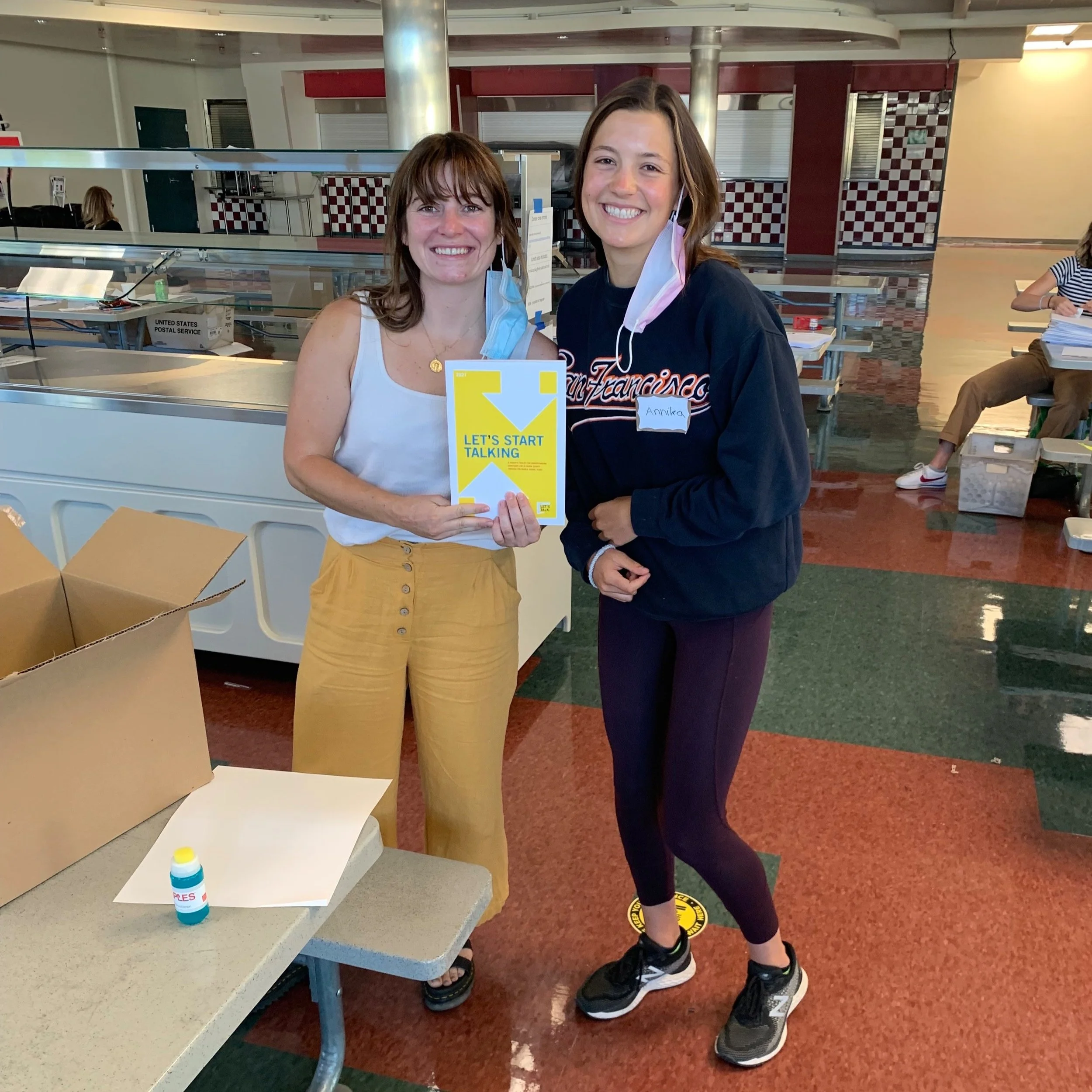 Two women standing inside an indoor space, smiling at the camera. The woman on the left is holding a yellow booklet titled 'Let's Start Talking,' and the woman on the right is wearing a dark sweatshirt, leggings, and sneakers. Masks are hanging off their ears.