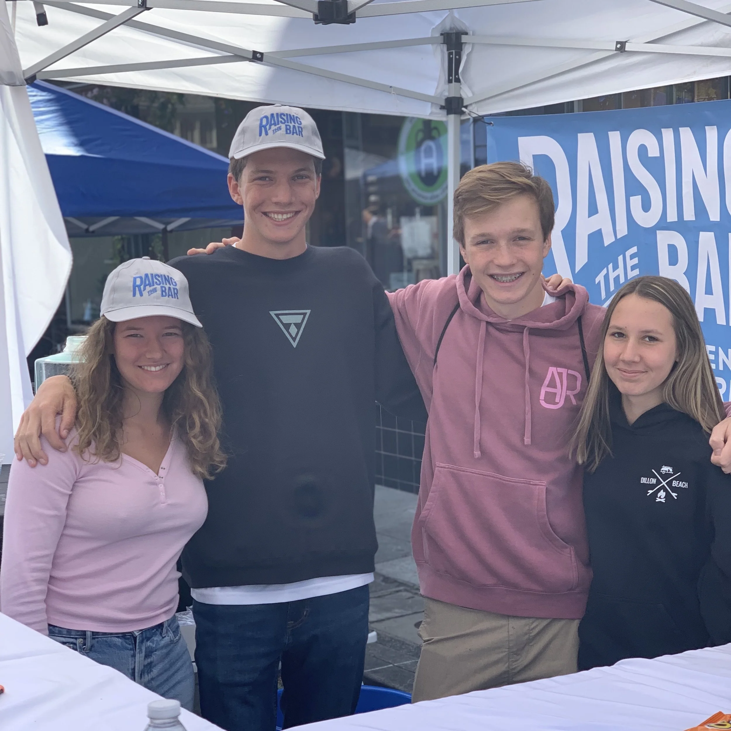 Group of four young people, three men and one woman, smiling and standing together at a Raising the Bar event under a white canopy tent with a blue banner in the background.