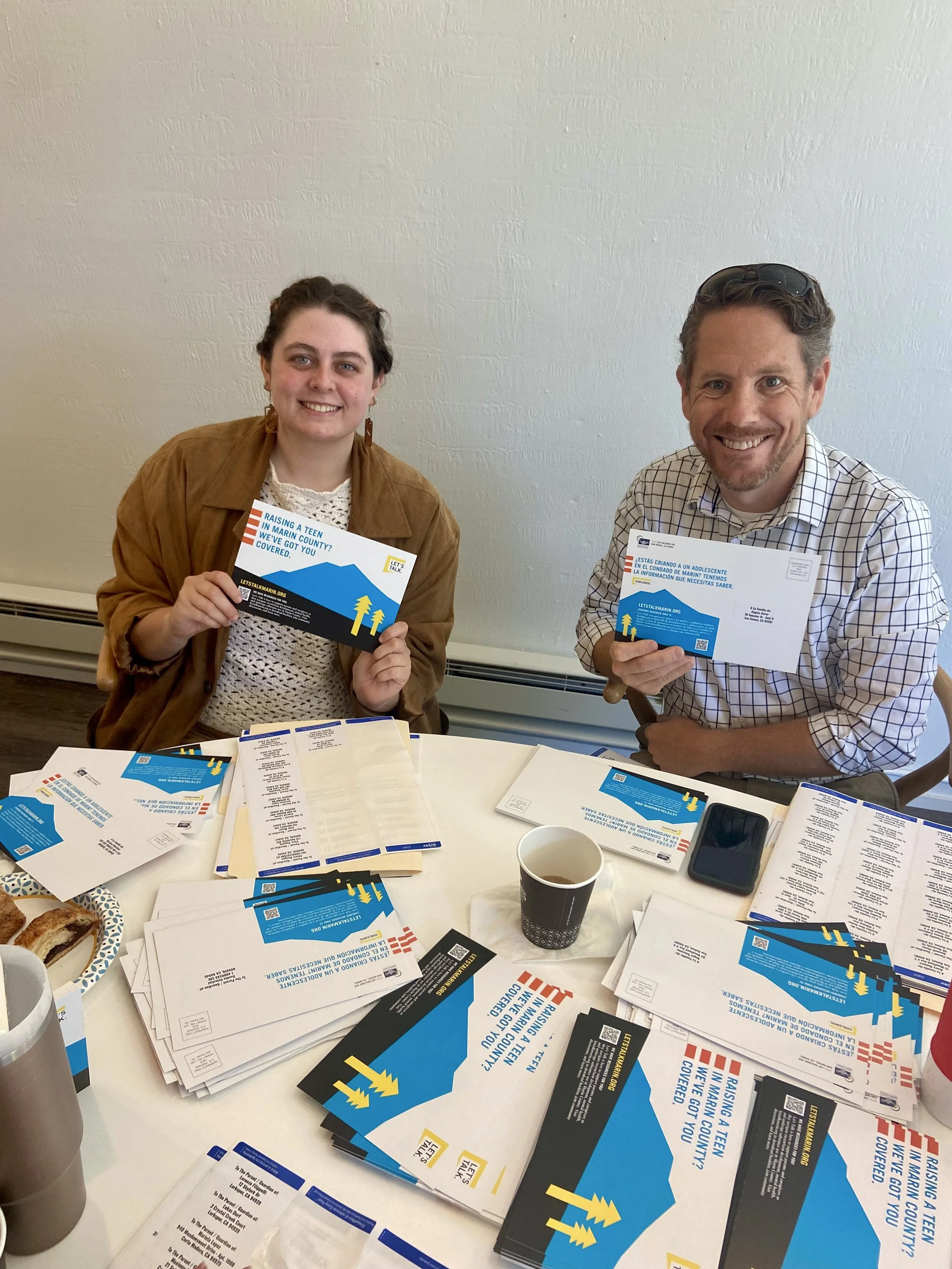 Two people sit at a table covered with flyers, papers, and cups, holding flyers promoting youth awareness in Maine. The woman on the left has brown hair, wears a brown jacket, and smiles at the camera. The man on the right has curly hair, sunglasses on his head, in a checkered shirt, also smiling.