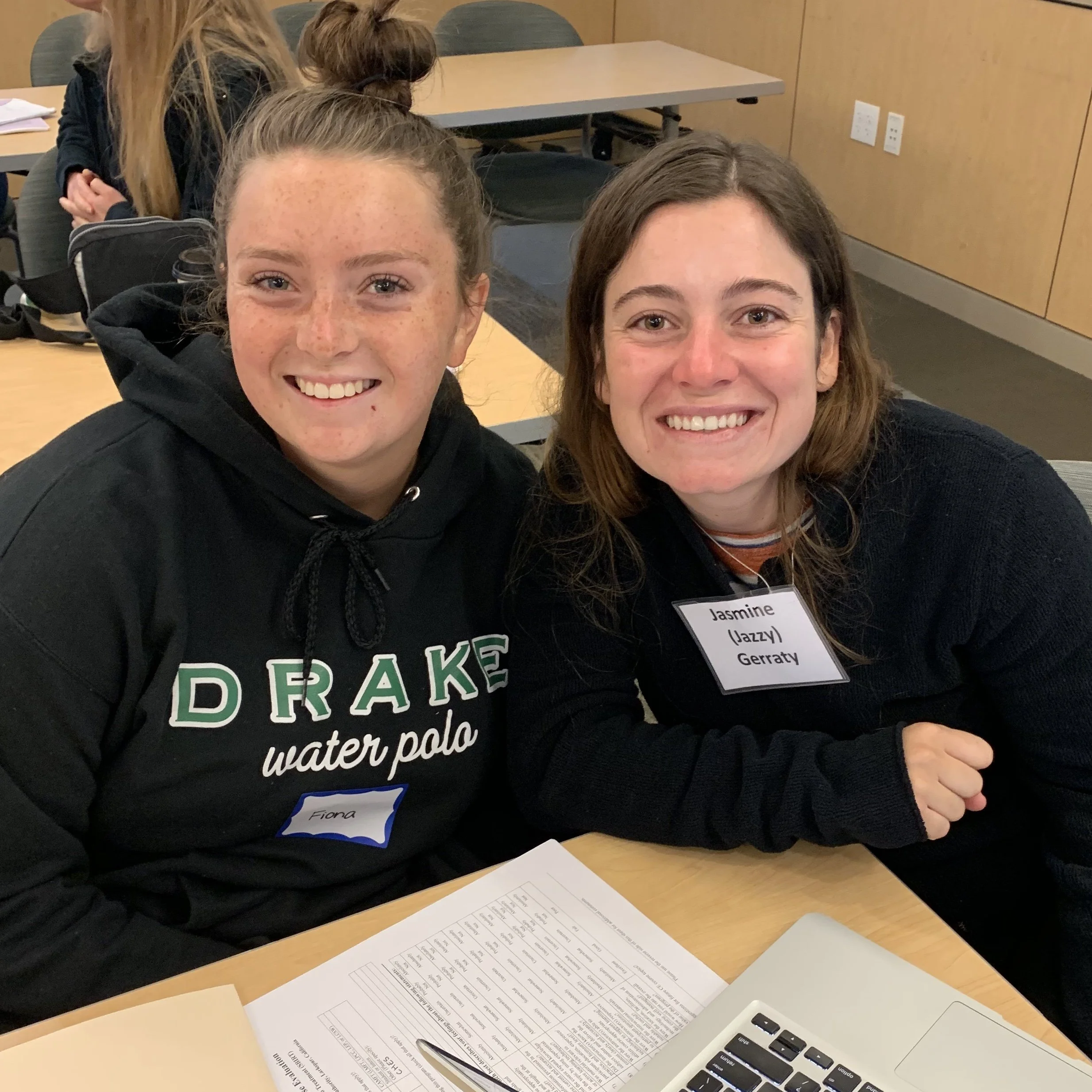 Two young women smiling at a table with papers and a laptop. The woman on the left wears a black hoodie with green and white text and a blue name tag. The woman on the right wears a black sweater with a name tag that reads Jasmine (Jazzy) Gerraty.